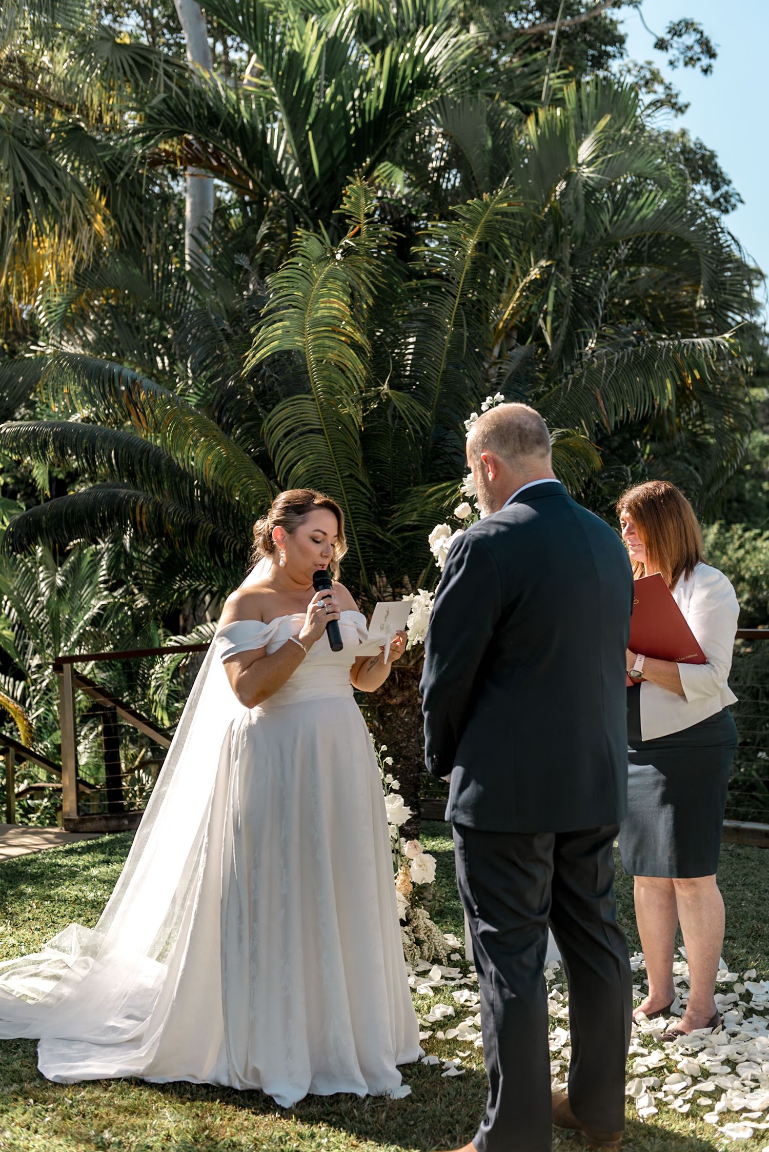 Bride smiles while reading vows into microphone at outdoor tropical wedding ceremony with lush green palms.