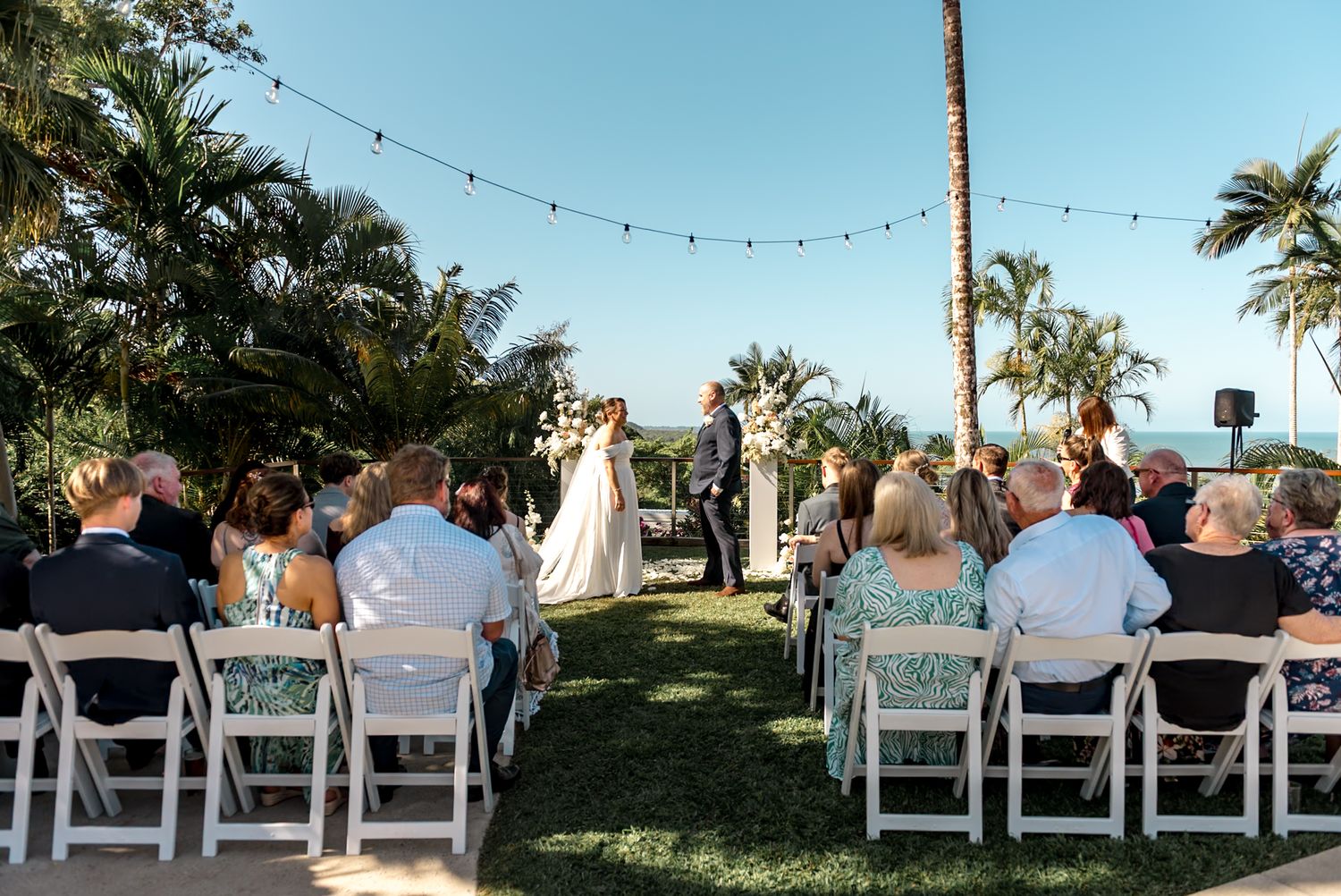 Guests seated in white chairs watch bride and groom exchange vows at outdoor tropical wedding ceremony.