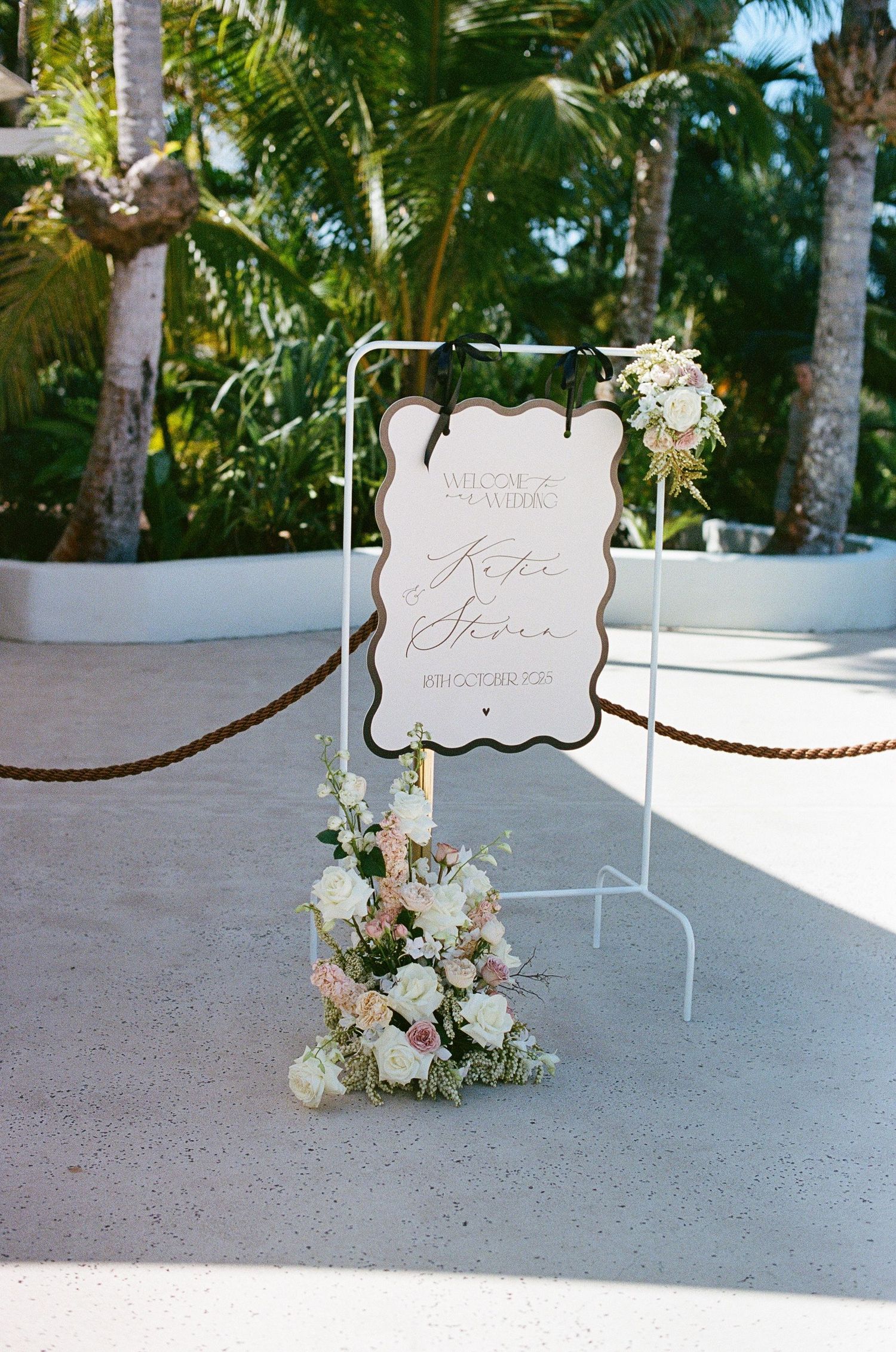 Elegant wedding welcome sign with floral decorations on a white stand surrounded by tropical palm trees.