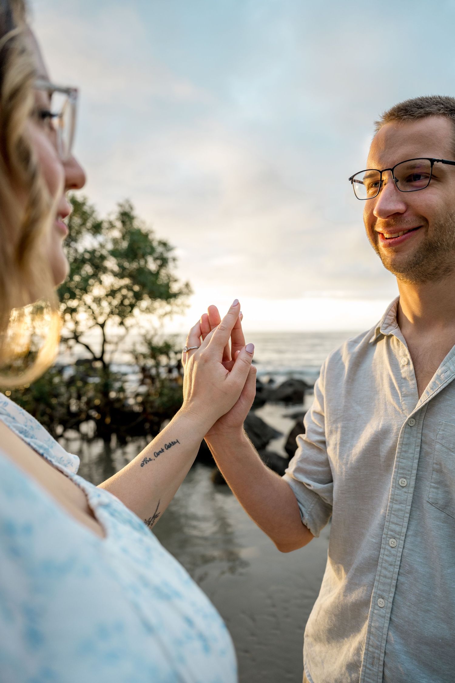 Smiling man with glasses holds hands with tattooed woman on sandy beach at golden sunset at Four Mile Beach, Port Douglas.