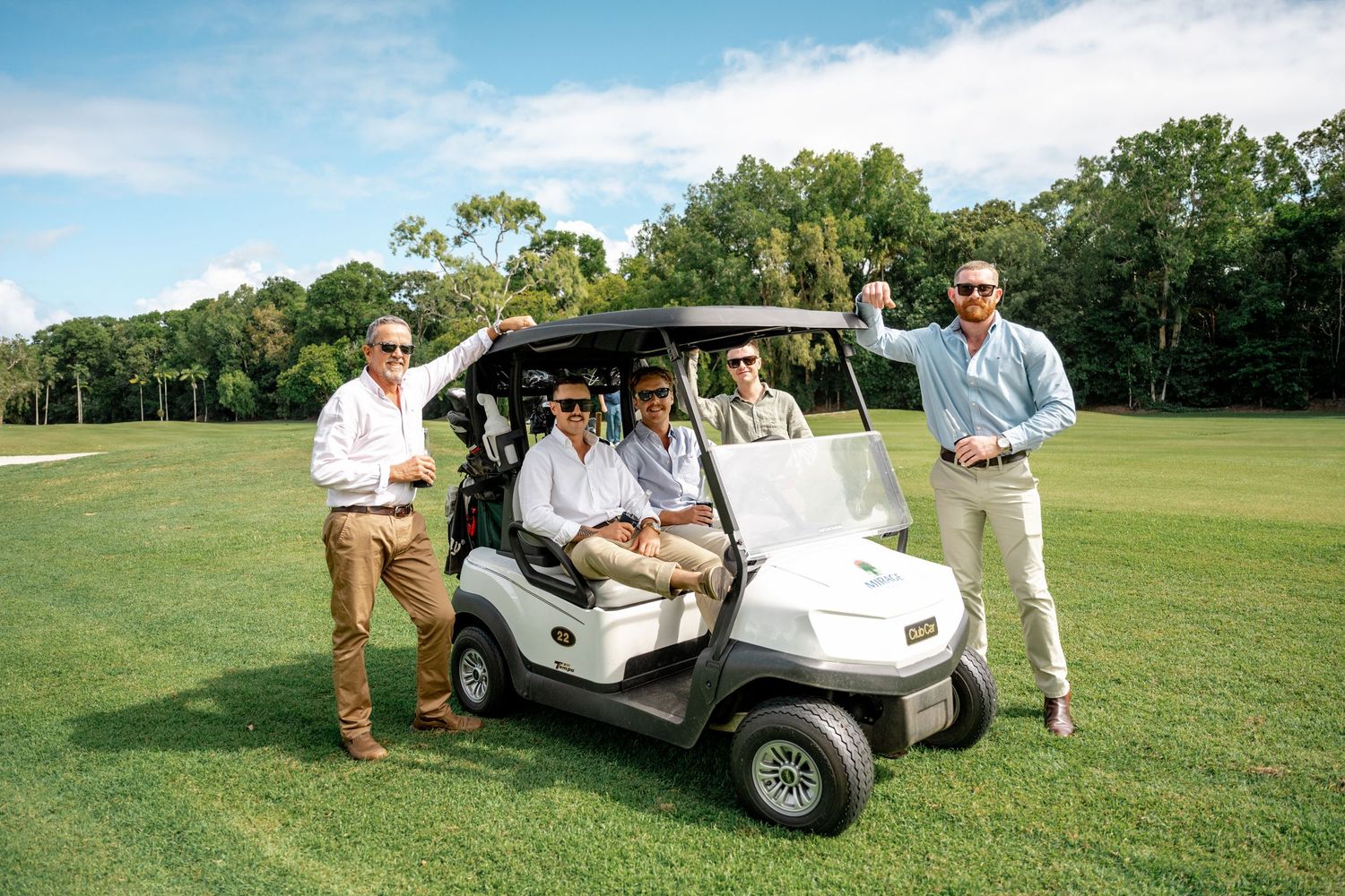 Group of five groomsmen posing with golf cart on lush green golf course under blue sky.