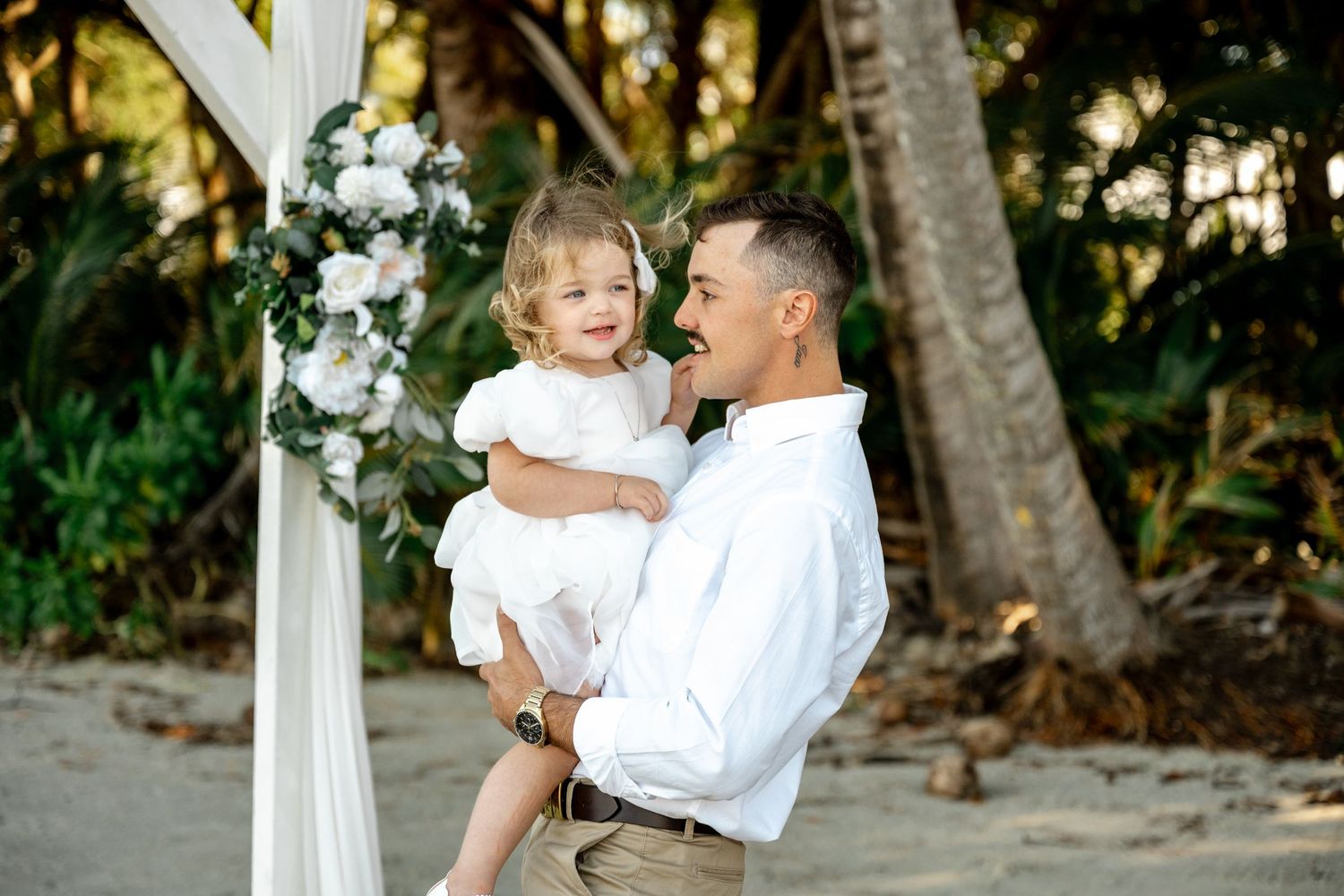 Smiling man in white shirt holds a happy toddler girl in white dress at a beach wedding arch at Four Mile Beach, Port Douglas.
