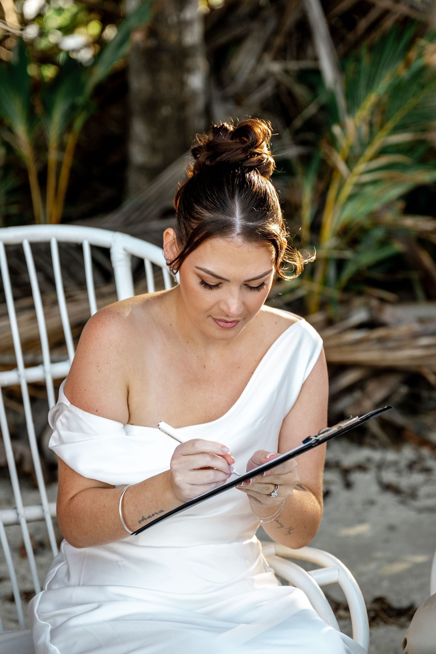 Beautiful bride in white off-shoulder gown signing marriage documents while seated on a white chair at Four Mile Beach, Port Douglas.
