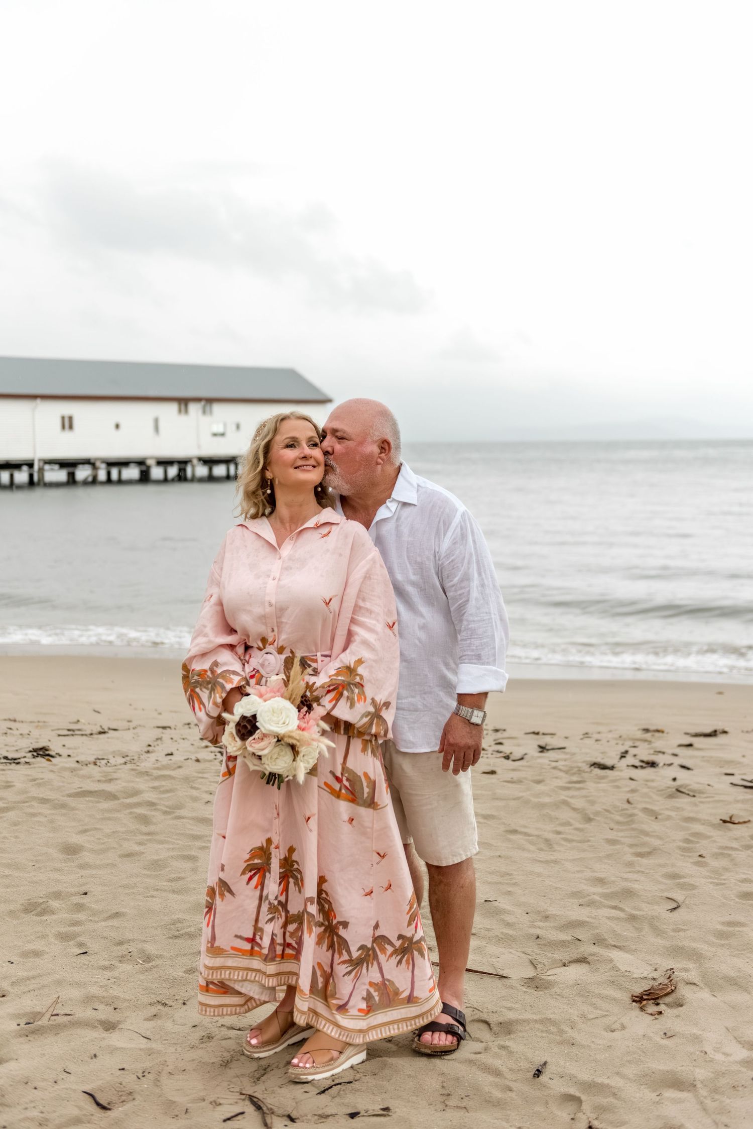 Couple sharing a romantic kiss on a sandy beach, bride holding a white floral bouquet wearing a pink palm print dress.
