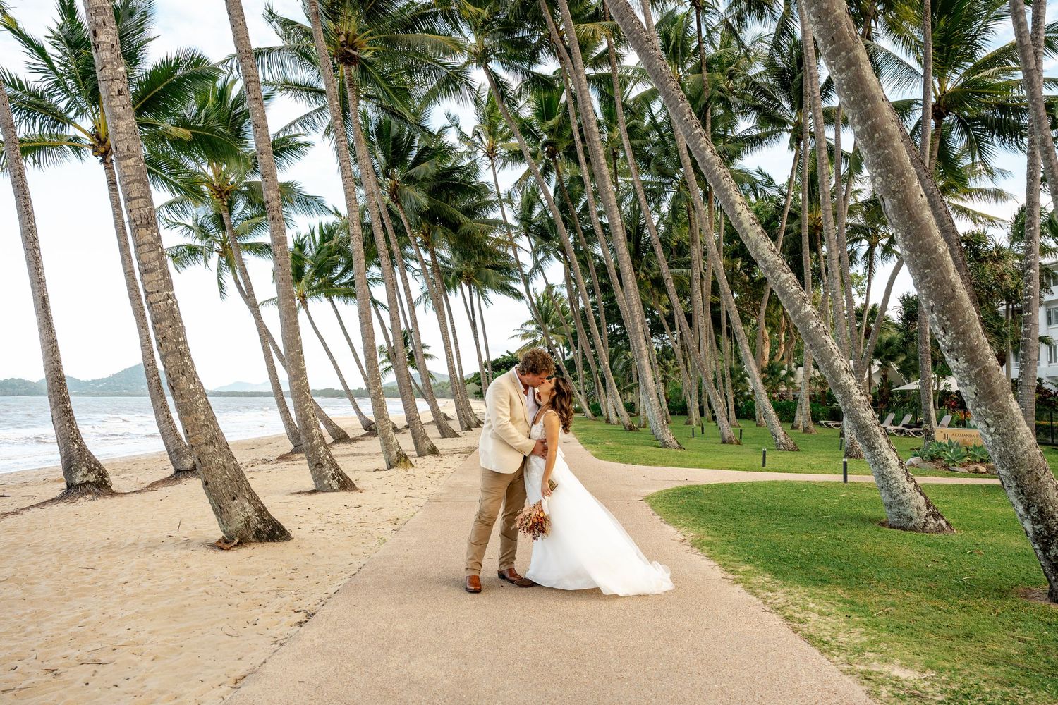 Bride and groom kissing on a palm-lined beach path with ocean in background.