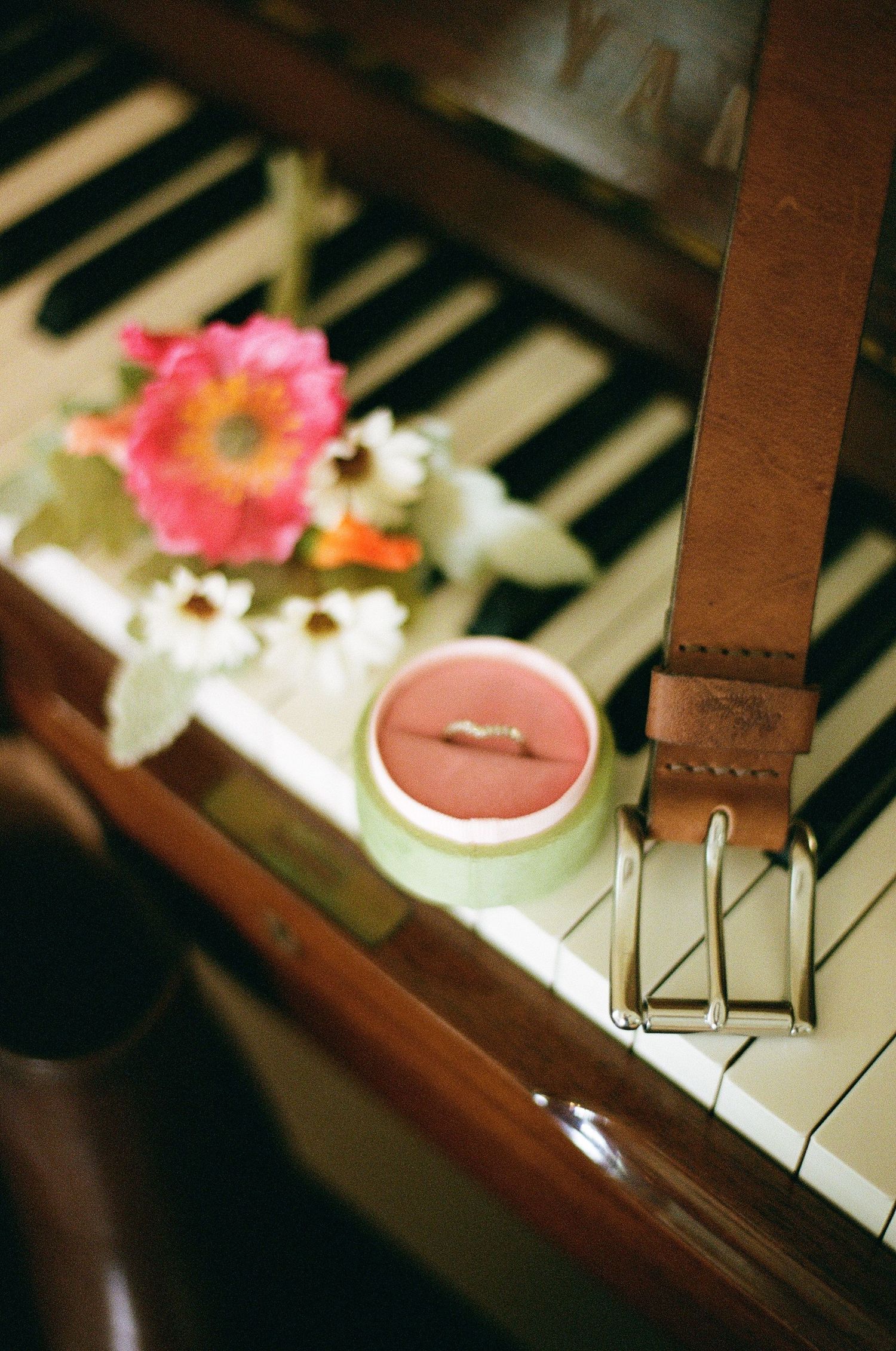 Wedding ring in pink box beside leather belt and flowers resting on vintage piano keys.
