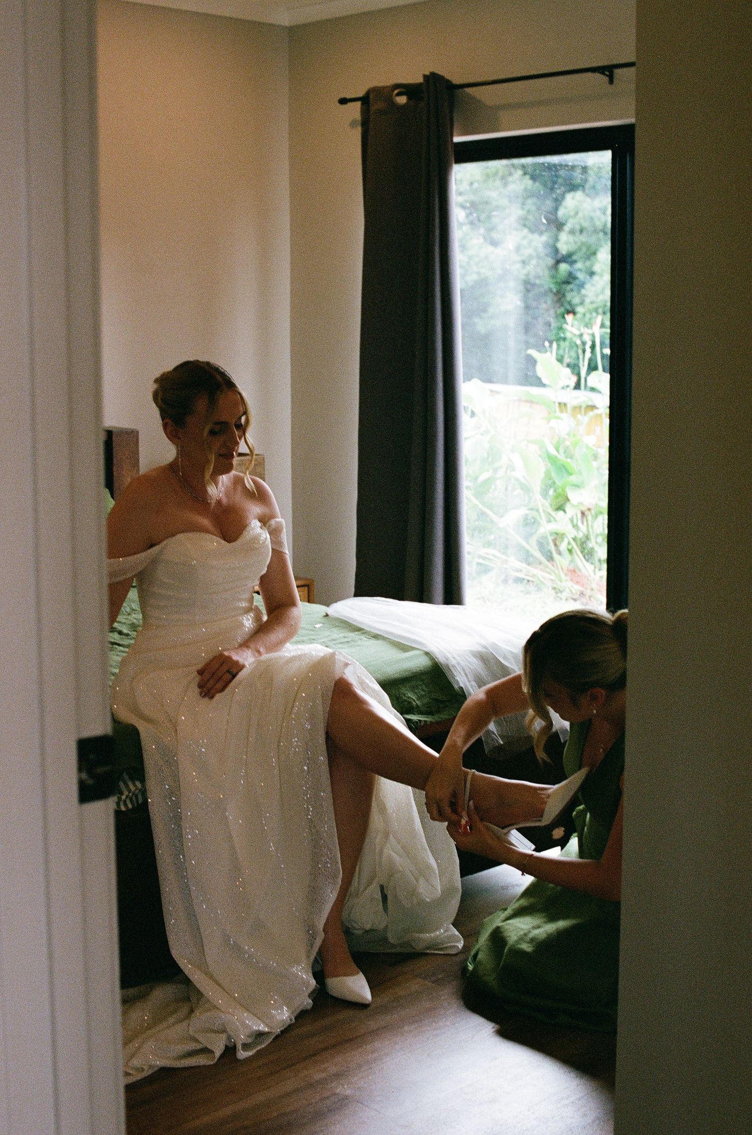 Bride in white strapless gown sits on bed while bridesmaid helps put on her wedding shoes.