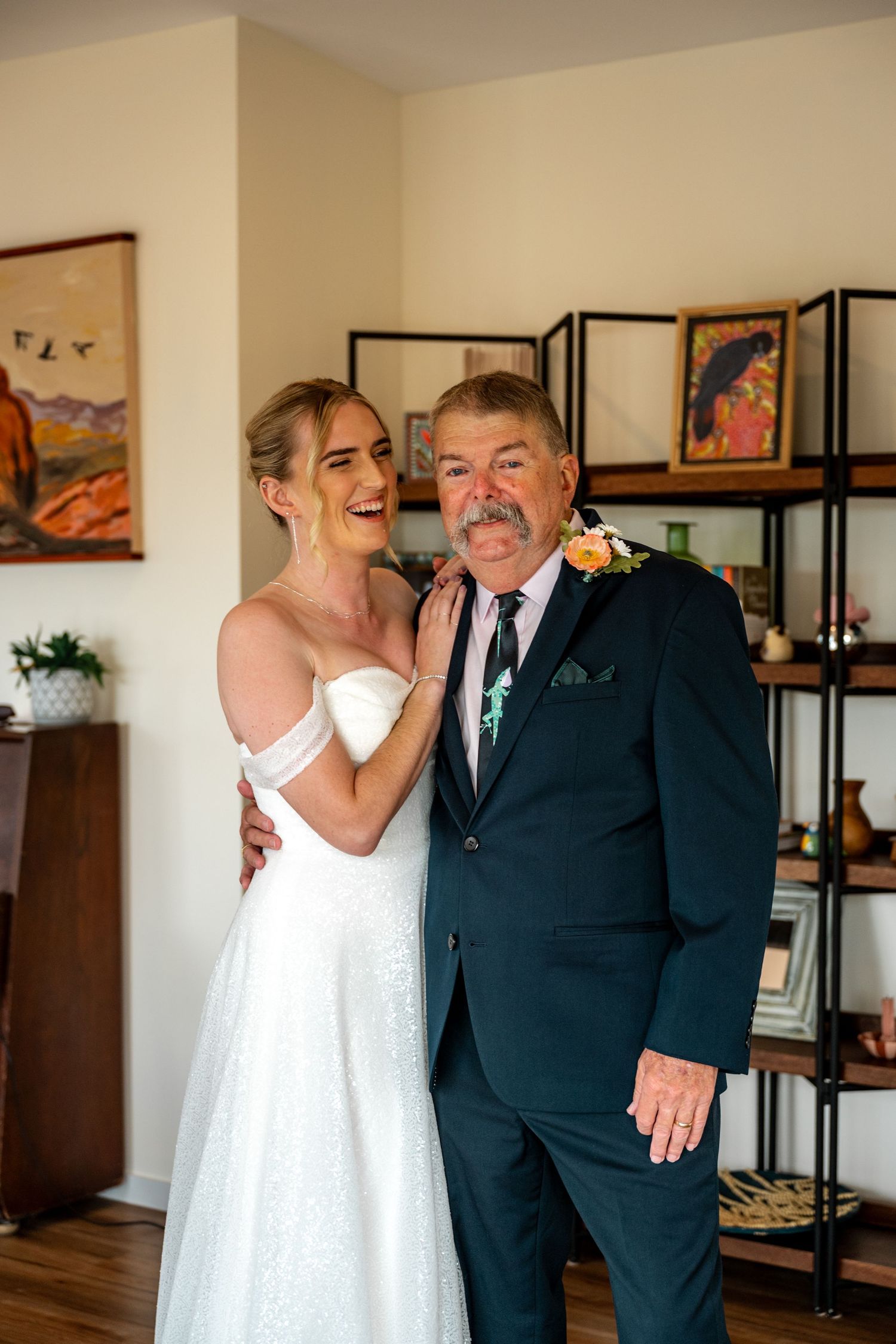 Laughing bride in white off-shoulder gown embracing father in navy suit before wedding.