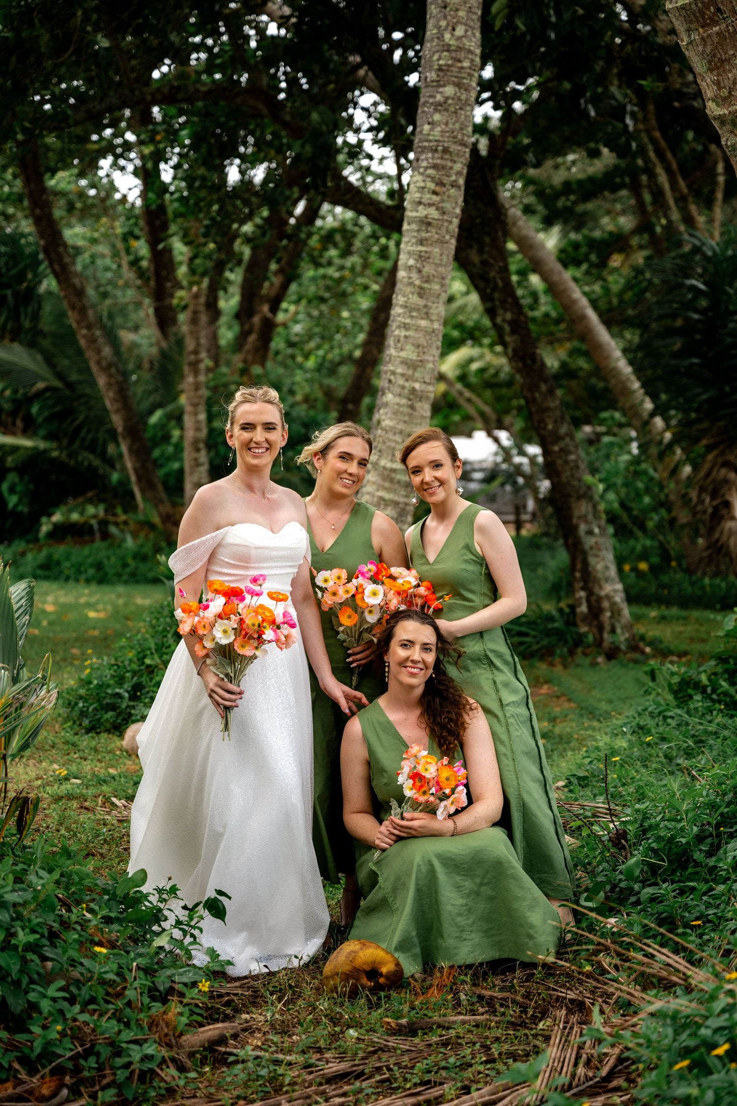 Bride in white off-shoulder gown poses with three bridesmaids in sage green dresses holding colorful bouquets in tropical garden.