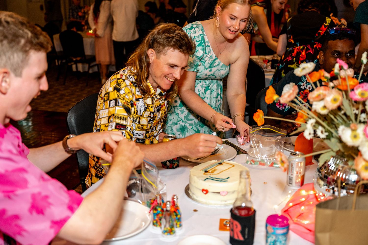 Wedding guests in colorful attire laughing while cutting and decorating white cake at reception table.
