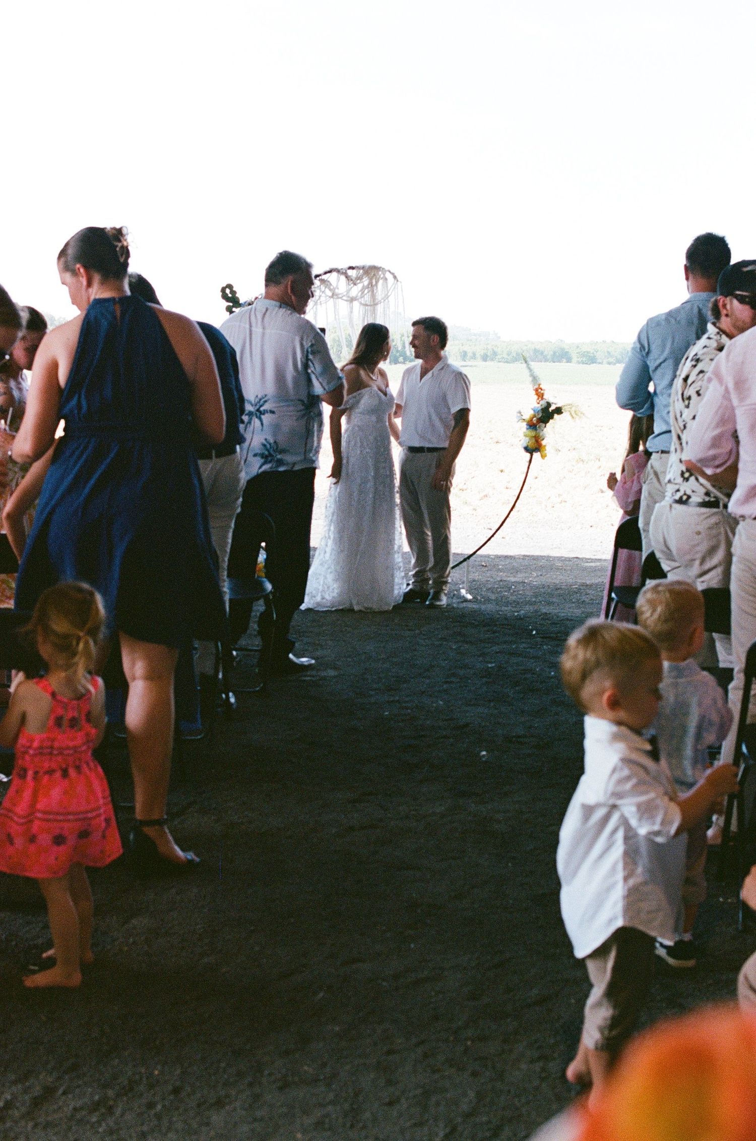 Outdoor beach wedding ceremony with bride in white lace gown, guests gathered on dark sand near waterfront.