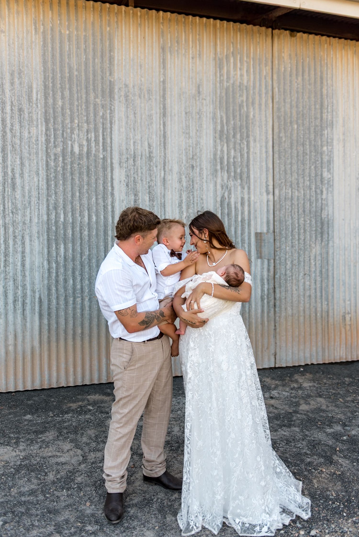 Bride and groom in wedding attire sharing tender moment with toddler and newborn baby outdoors.