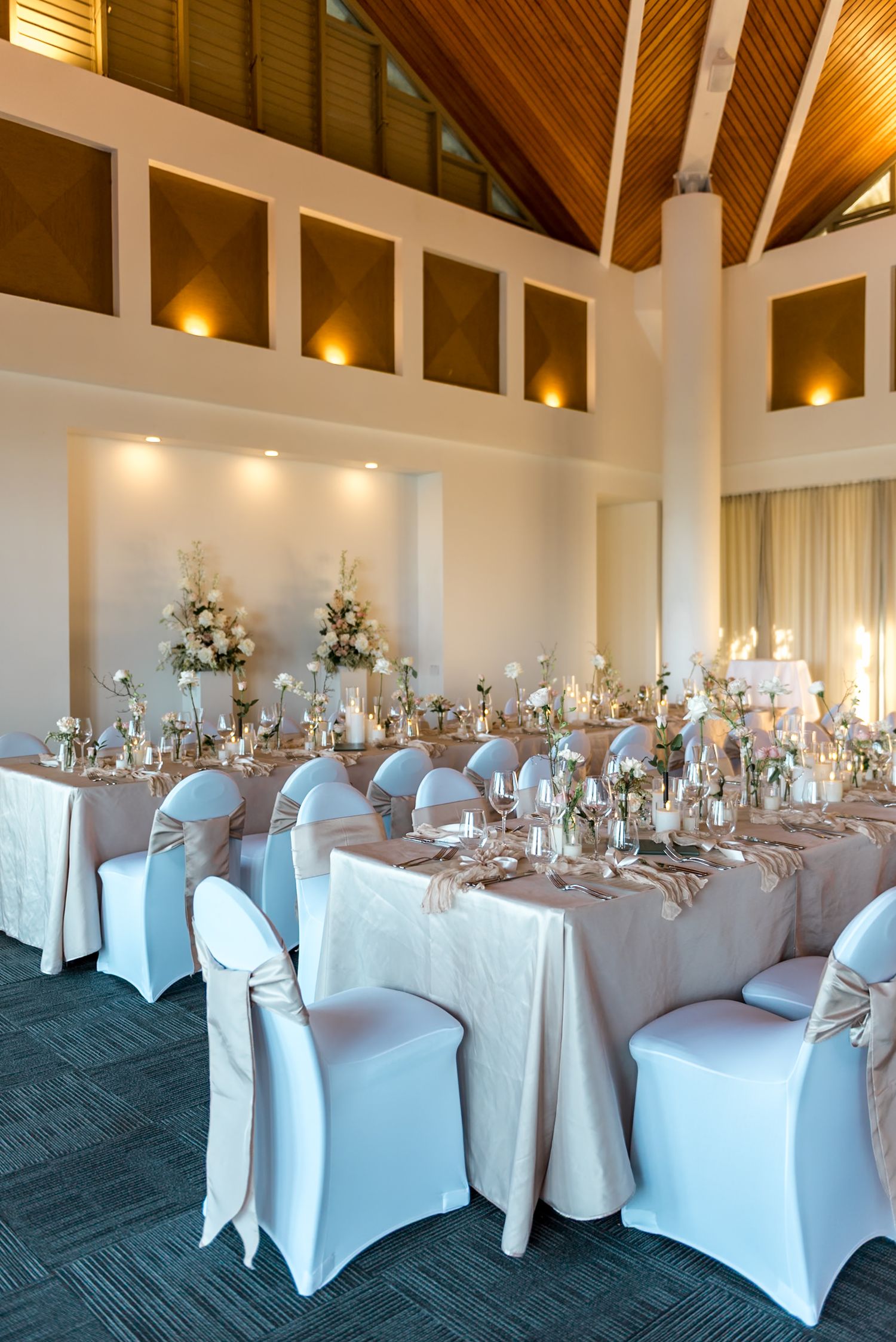 Luxurious wedding banquet hall with high timber ceiling, white chair covers, and floral centerpieces on long tables.