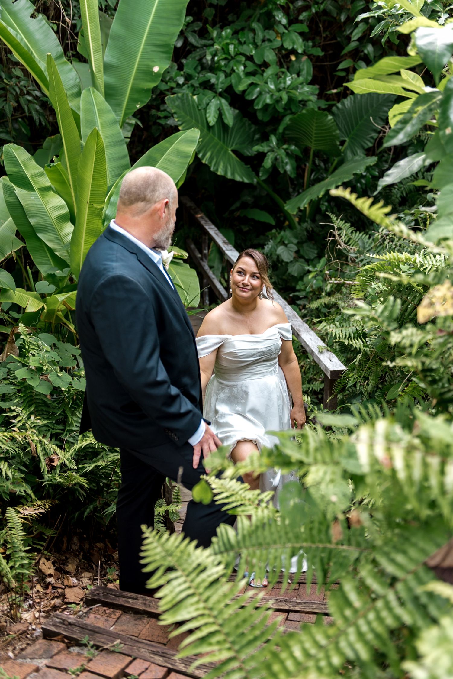 Smiling bride in short white off-shoulder dress climbing steps toward groom in tropical garden.