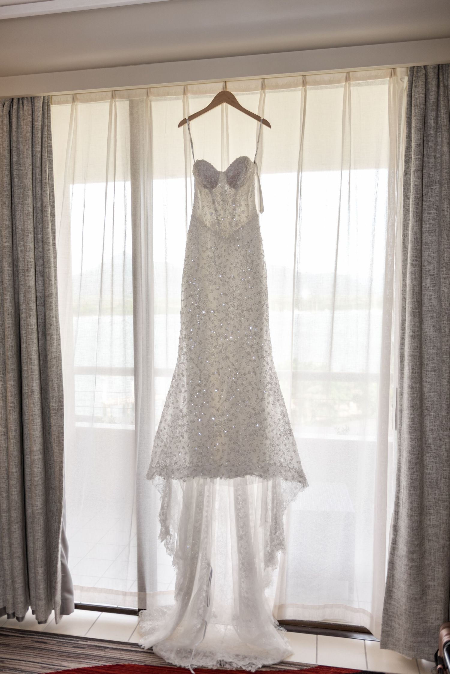 Elegant strapless beaded mermaid wedding gown hanging on a wooden hanger in front of sheer curtained window.