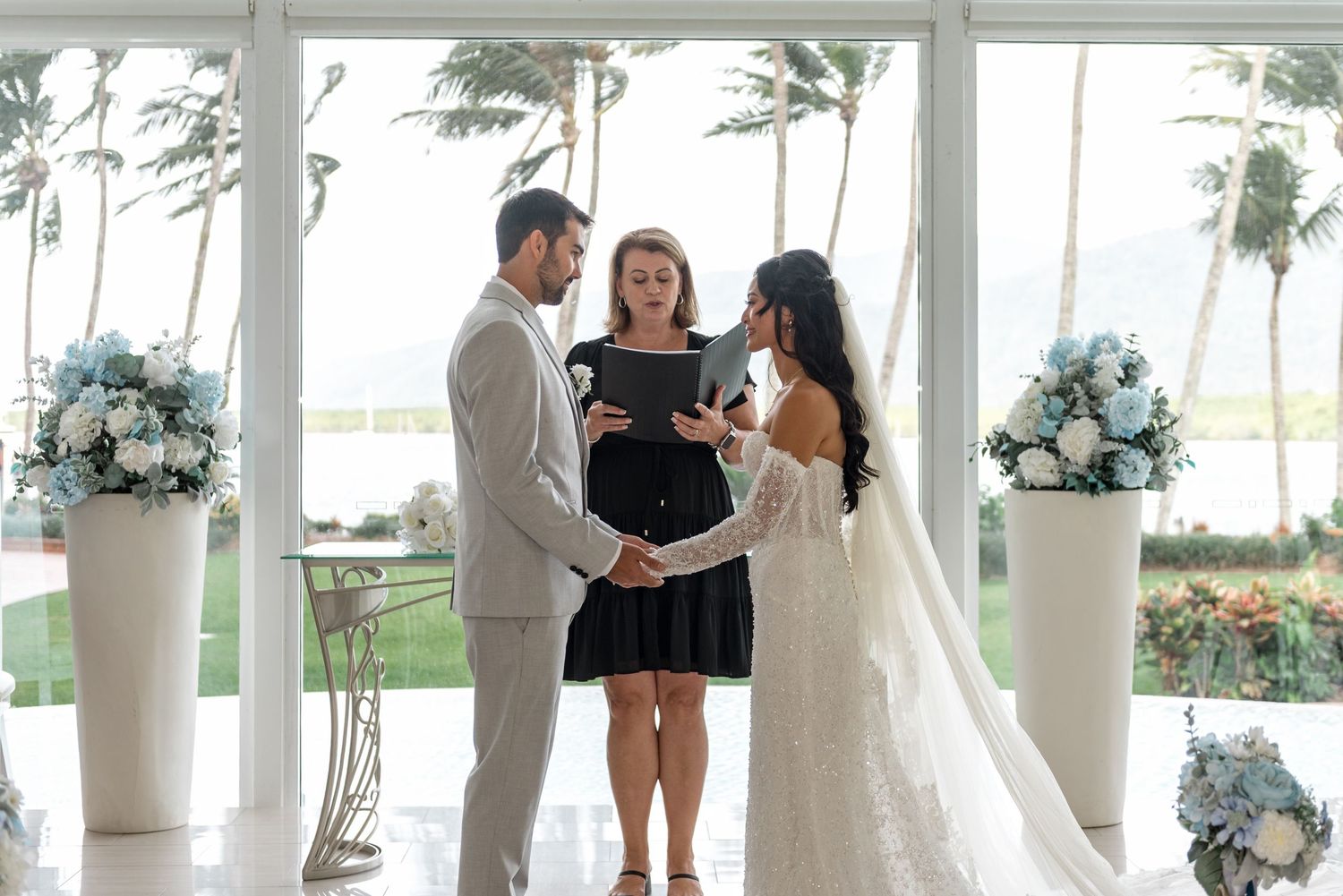 Bride and groom exchange vows with celebrant in tropical beachside glass chapel.