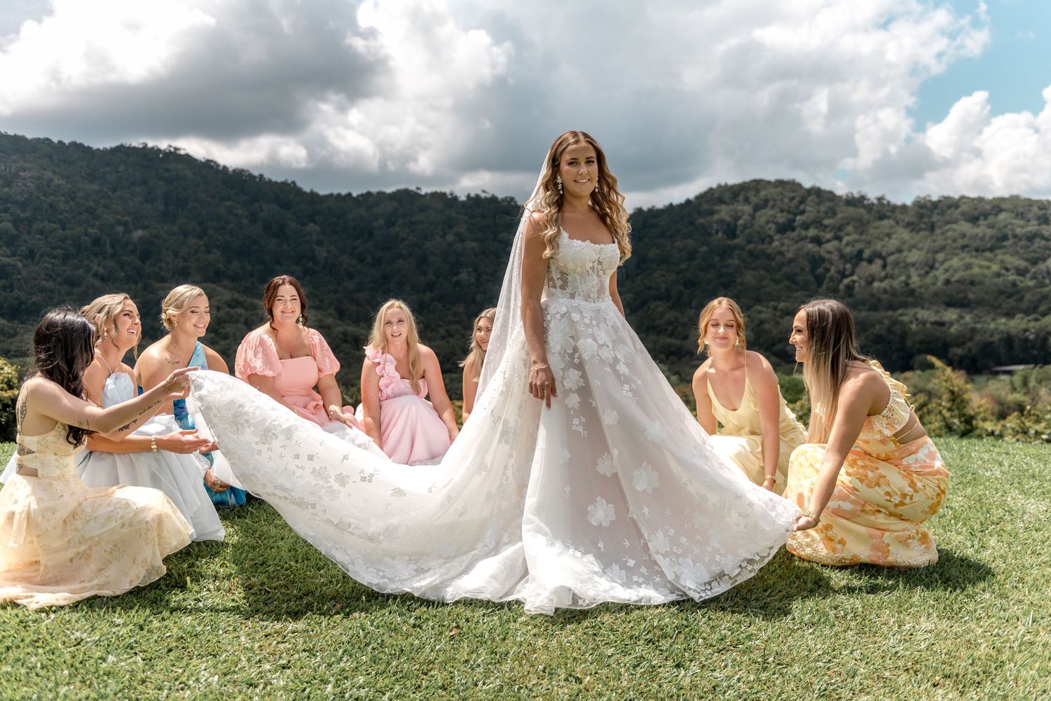 Bridesmaids spread elegant lace wedding gown train outdoors with lush mountain scenery behind.