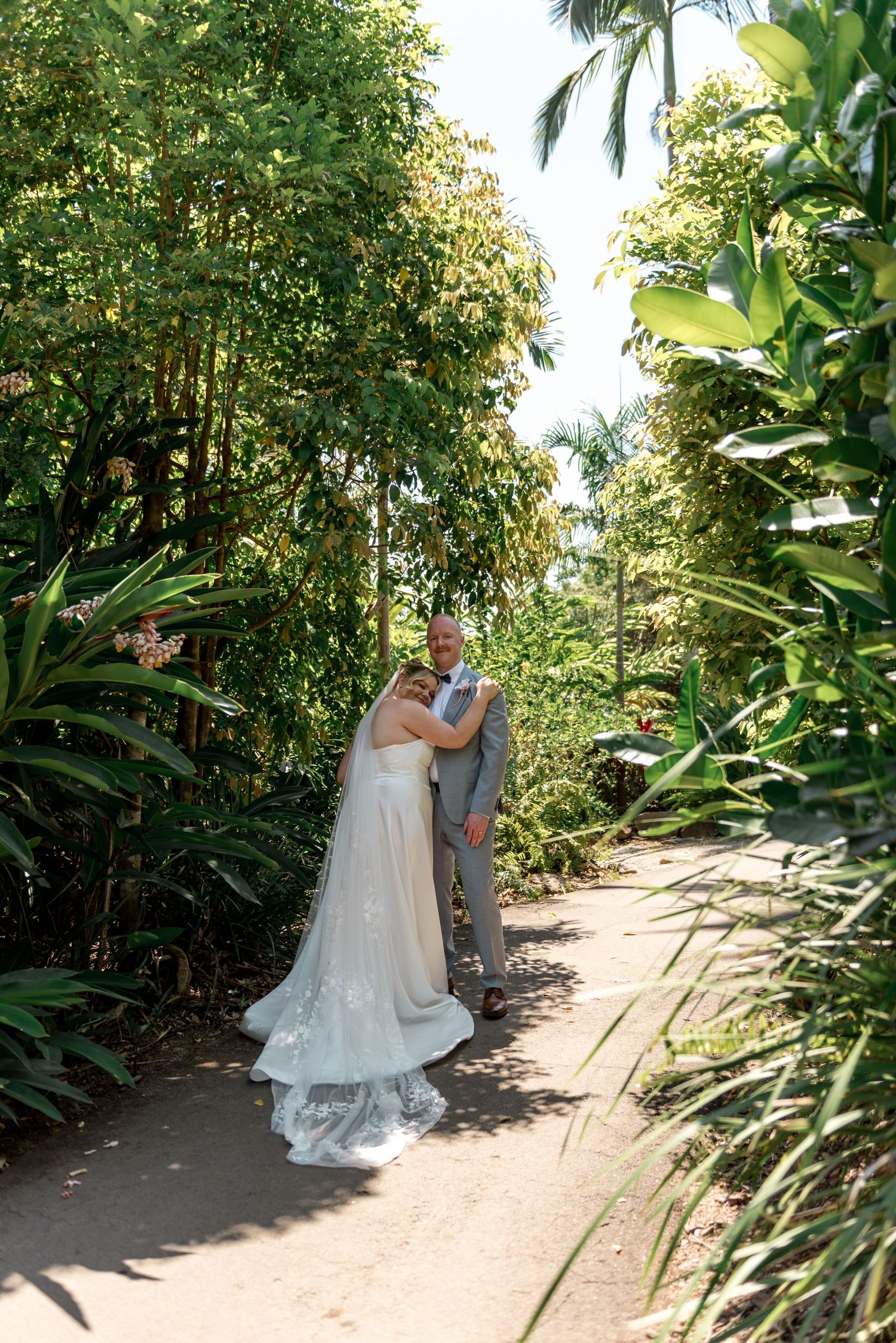 Bride and groom pose together on a sunlit tropical garden path surrounded by vibrant green palm foliage.