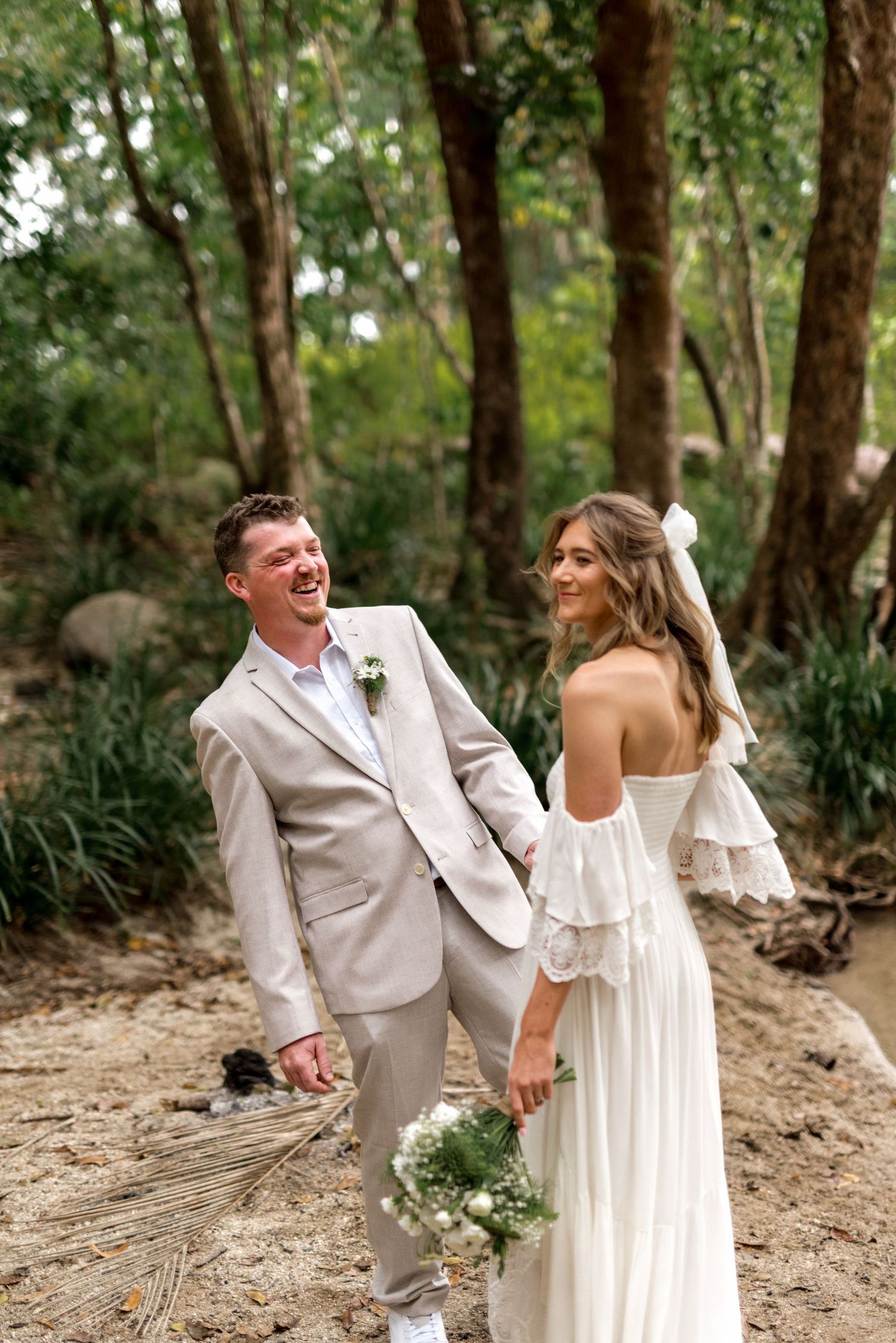 Laughing bride and groom hold hands in a lush forest during their wedding shoot.