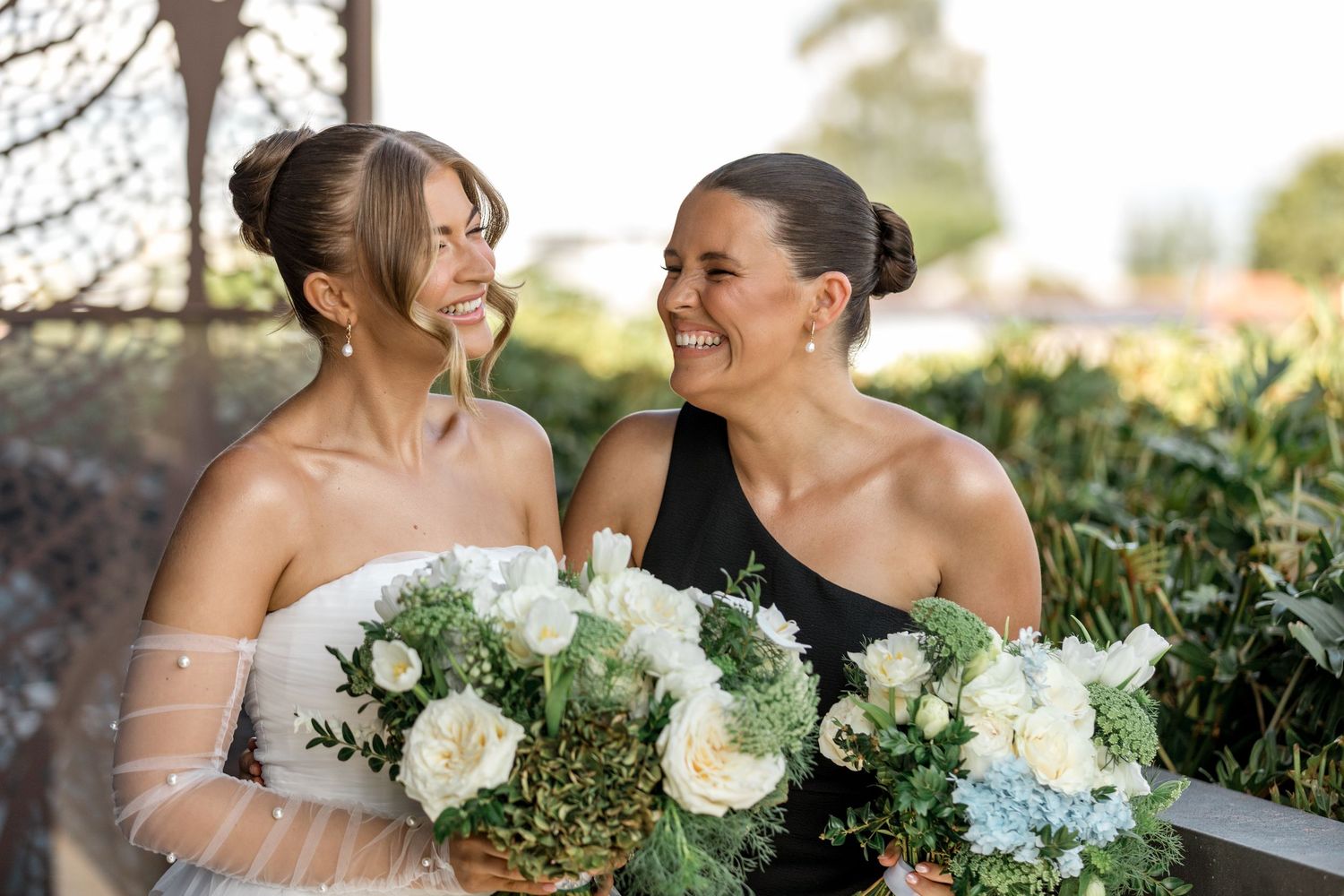Bride in white gown and bridesmaid in black dress laugh together holding white floral bouquets outdoors.