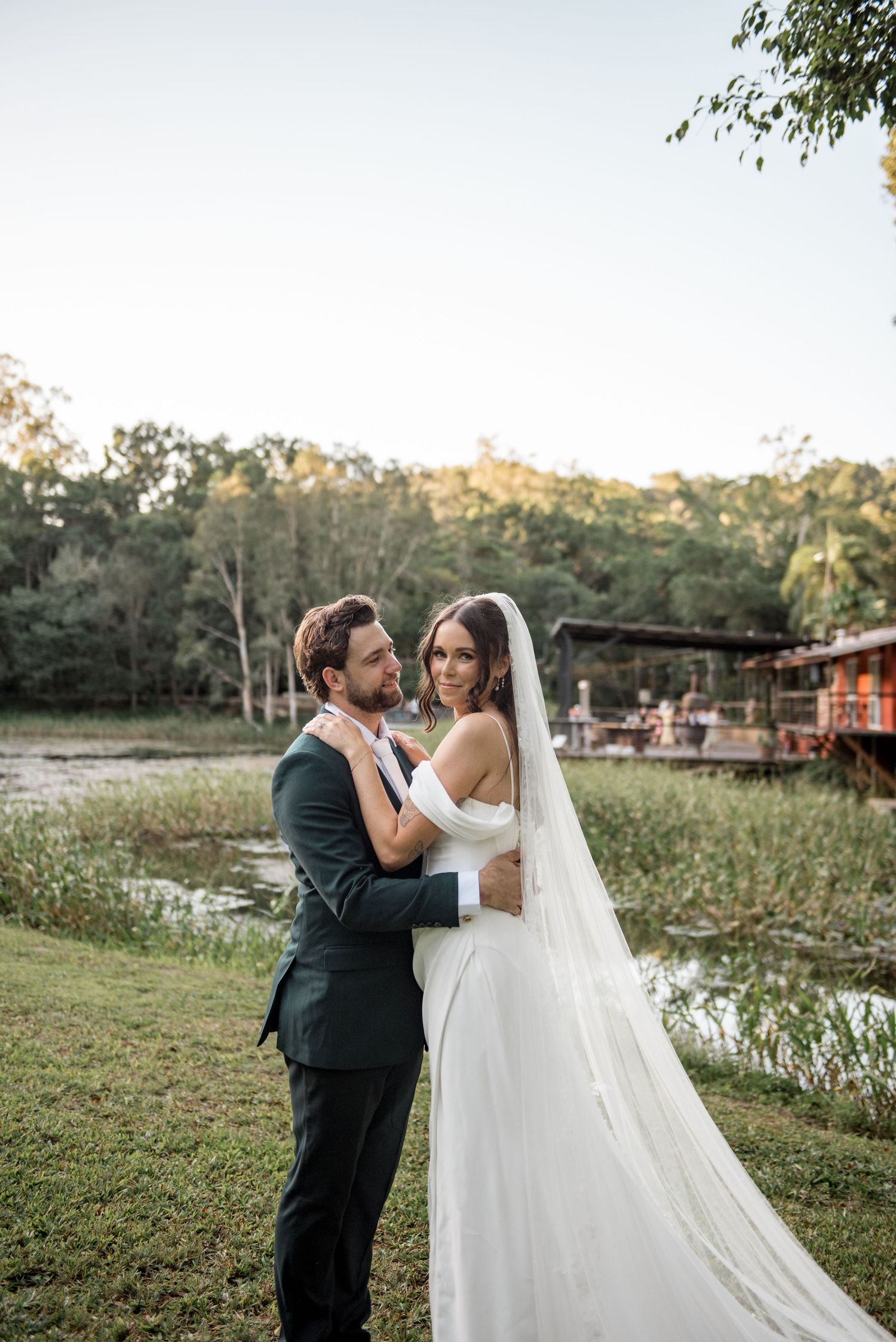 Bride in off-shoulder gown with cathedral veil embraces groom in dark suit by a tranquil pond.