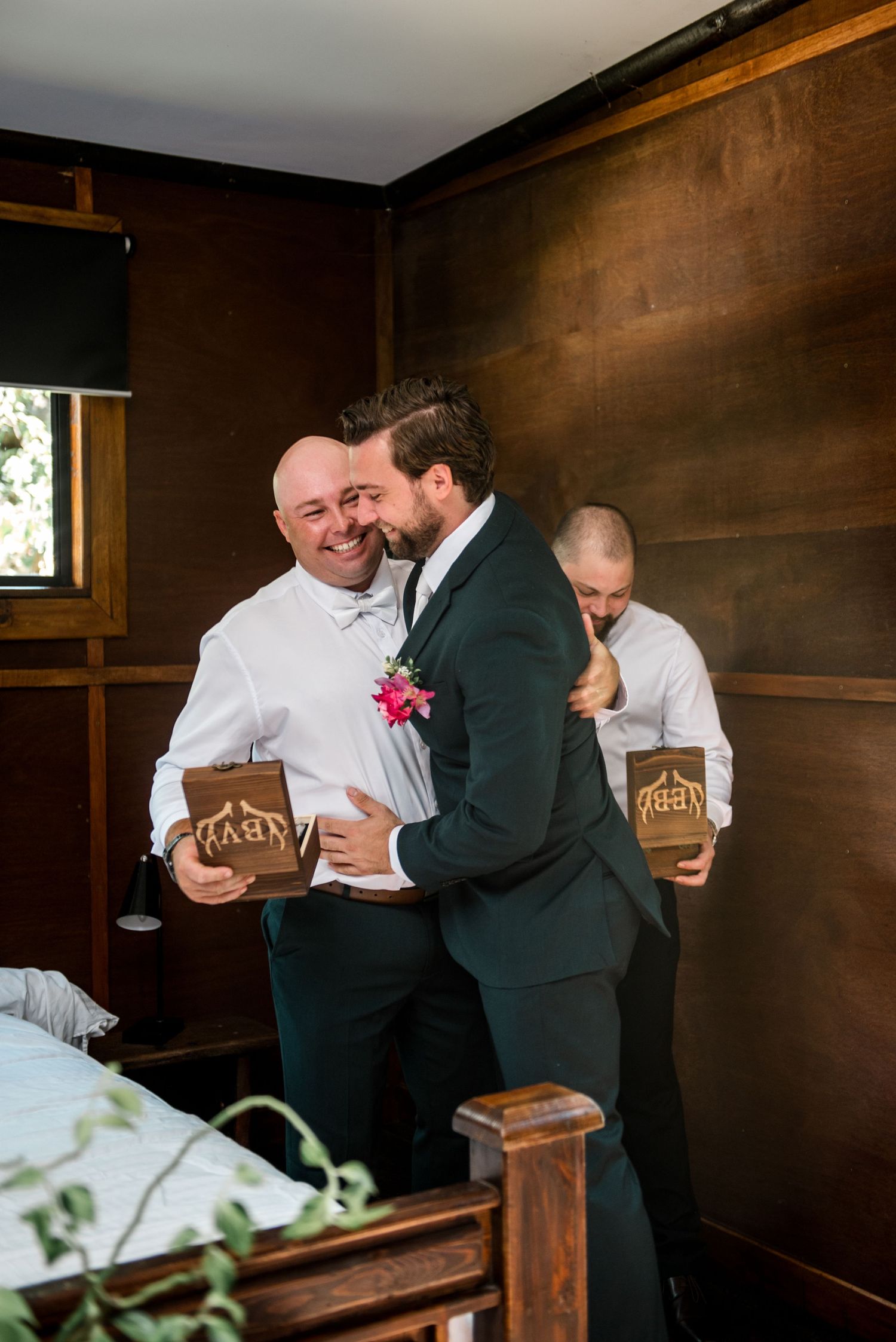 Two groomsmen share a warm hug while holding wooden gift boxes during wedding preparations indoors.