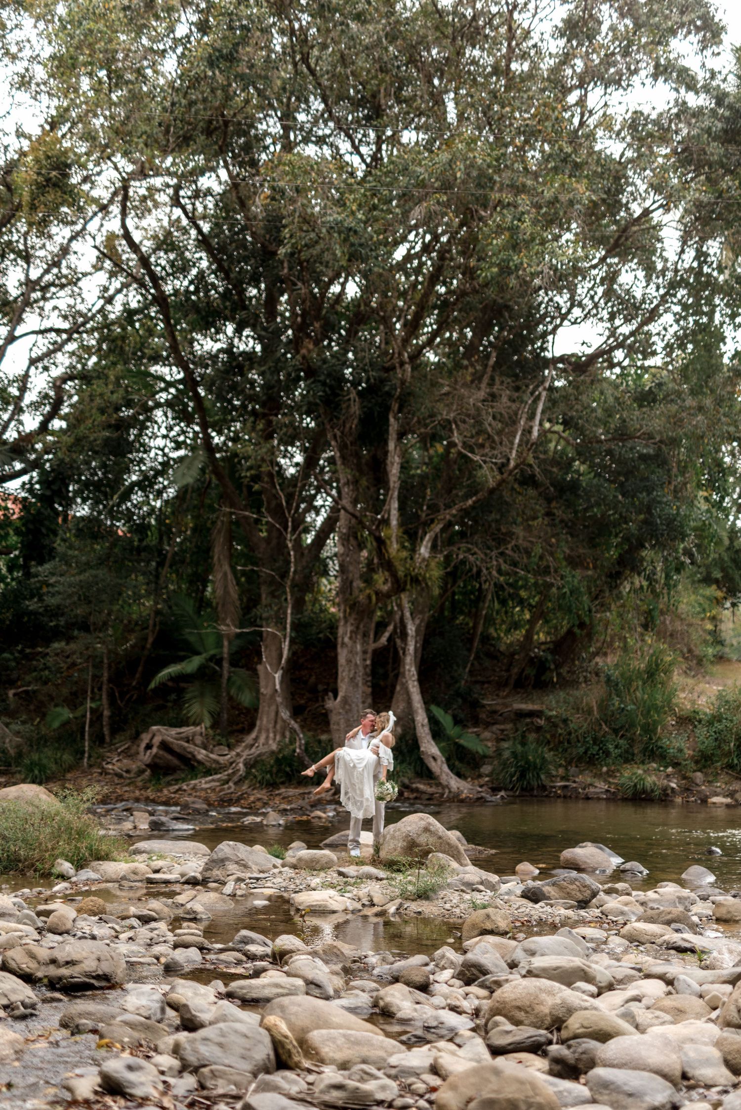 Bride and groom stand on rocky riverbank surrounded by lush green trees during outdoor wedding photo session.
