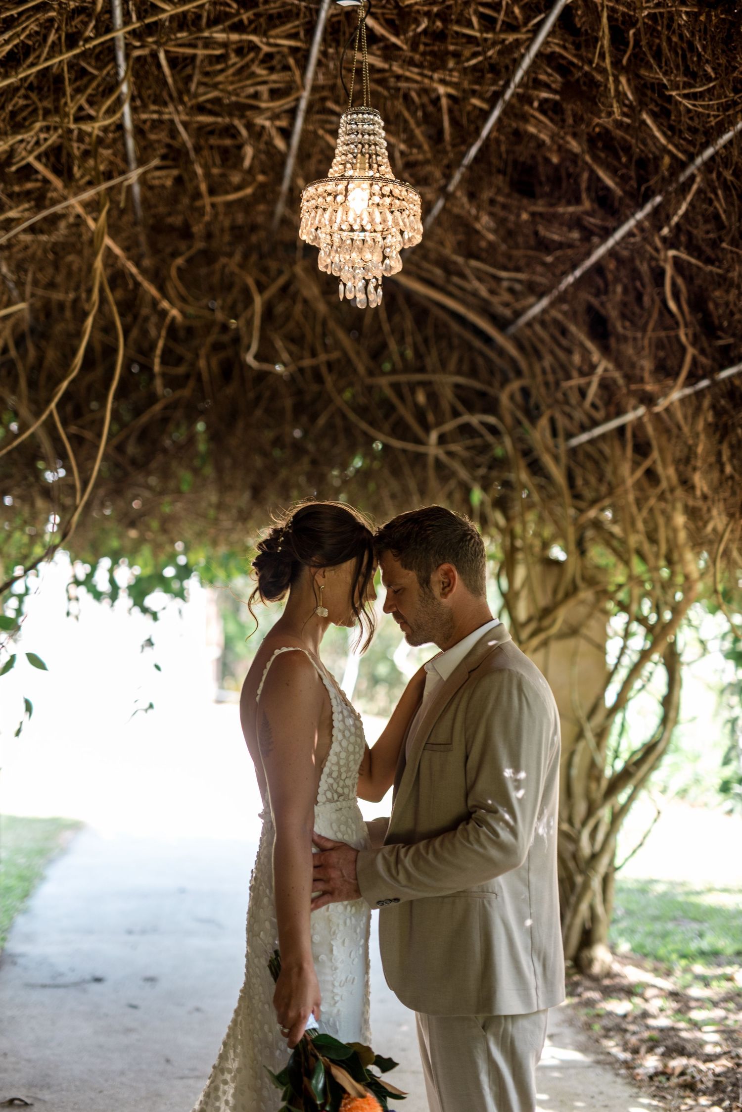 Bride and groom touch foreheads romantically beneath a crystal chandelier in a vine tunnel.