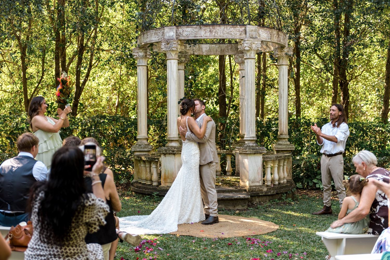 Newlyweds share first kiss at stone gazebo altar as guests celebrate their outdoor garden wedding ceremony.