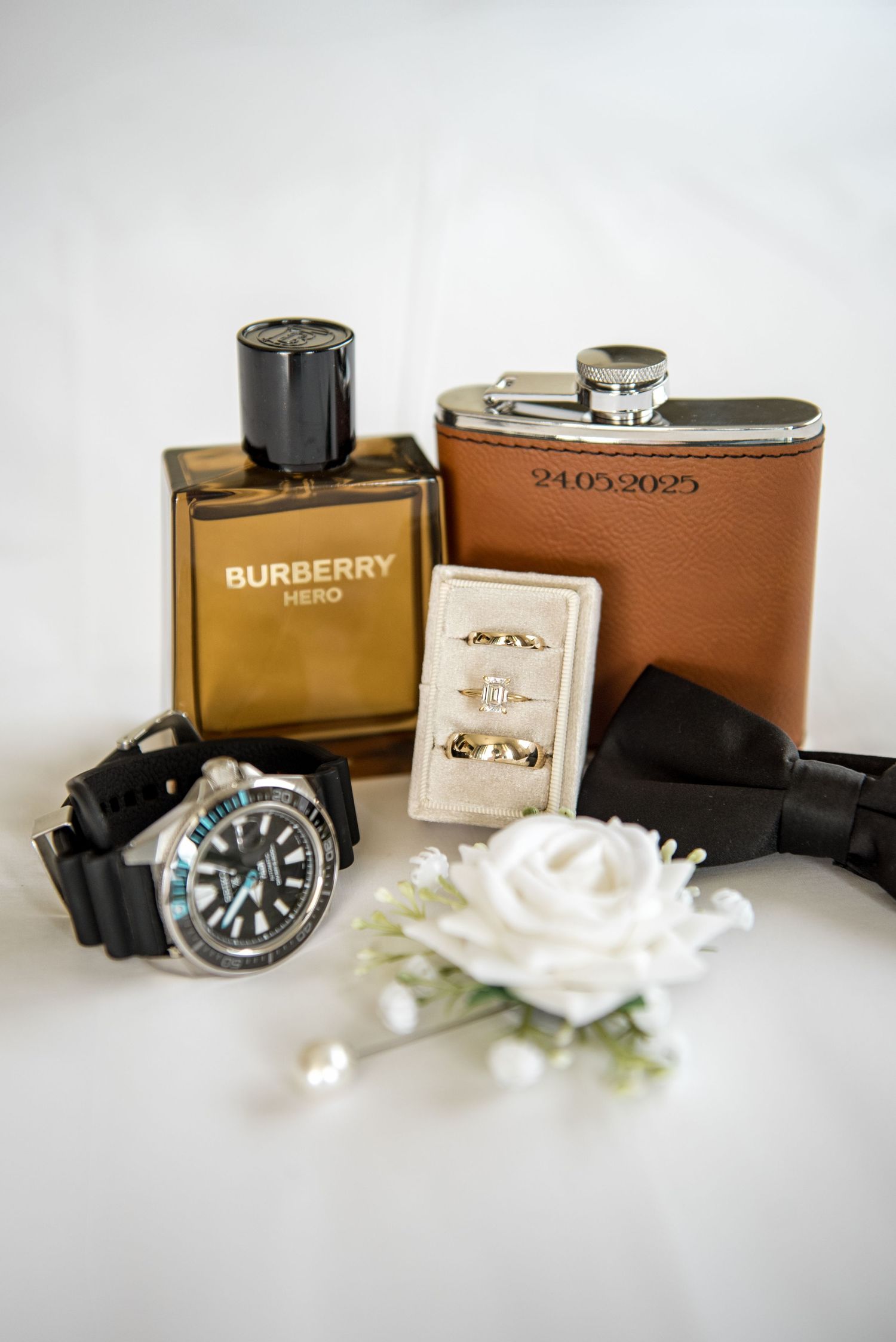 Wedding flat lay featuring Burberry Hero cologne, a engraved flask dated 24.05.2025, watch, rings, and boutonniere.