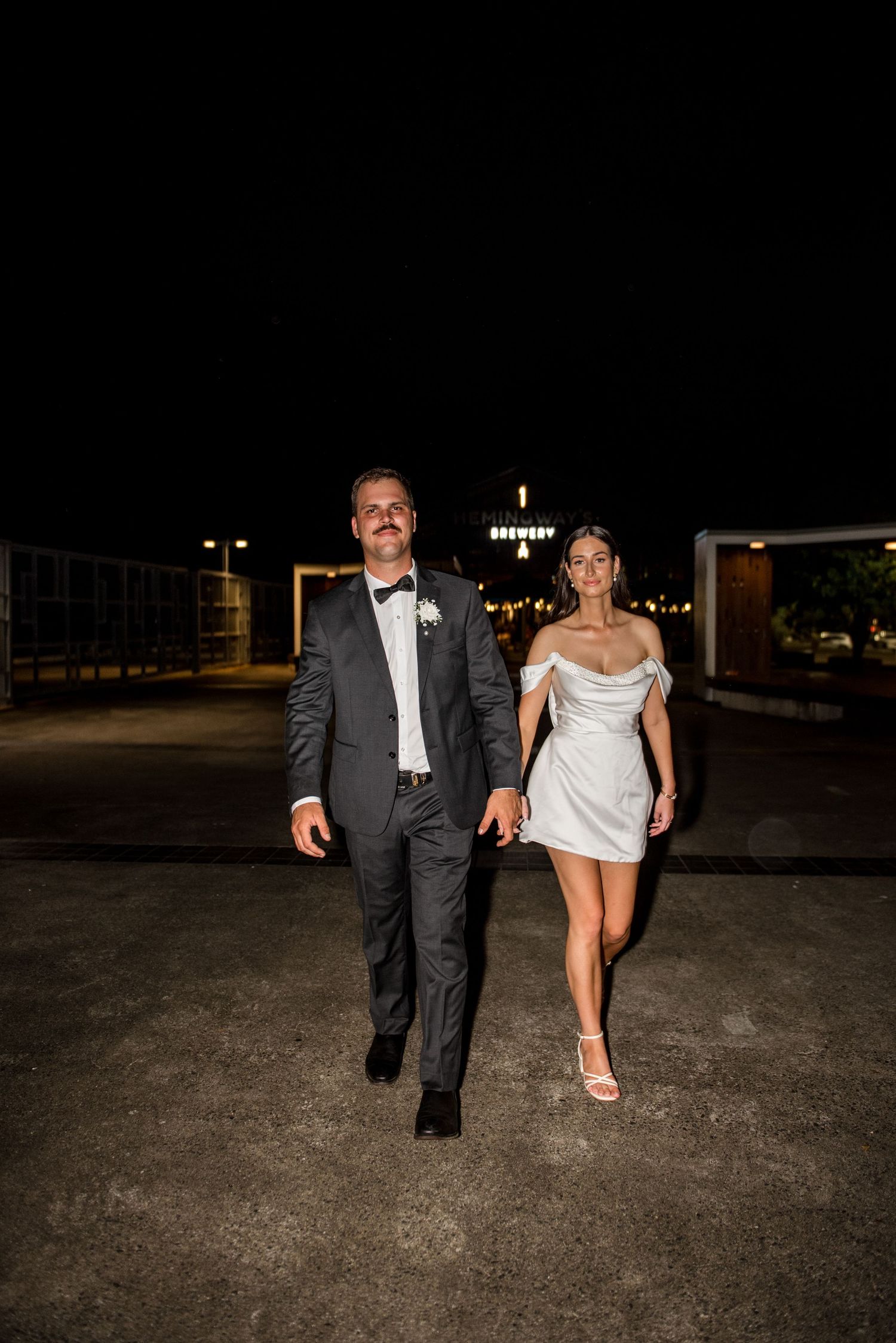 Newlyweds walk hand in hand outdoors at night near Hemingways Brewery after their wedding.