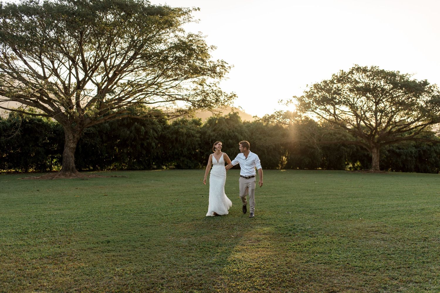 Bride and groom walking hand in hand across green lawn at golden sunset.