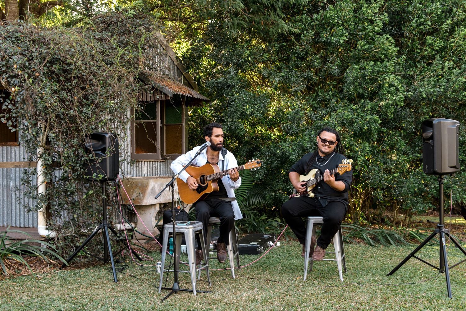 Two musicians play acoustic guitars outdoors at a wedding reception in a garden setting.
