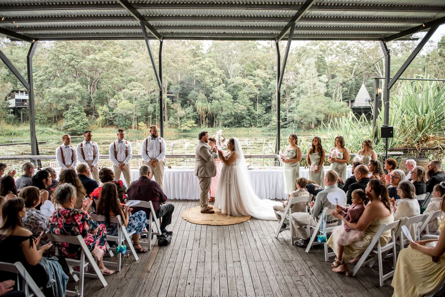 Outdoor wedding ceremony with bride and groom at altar, guests seated on white chairs, lush green trees in background.
