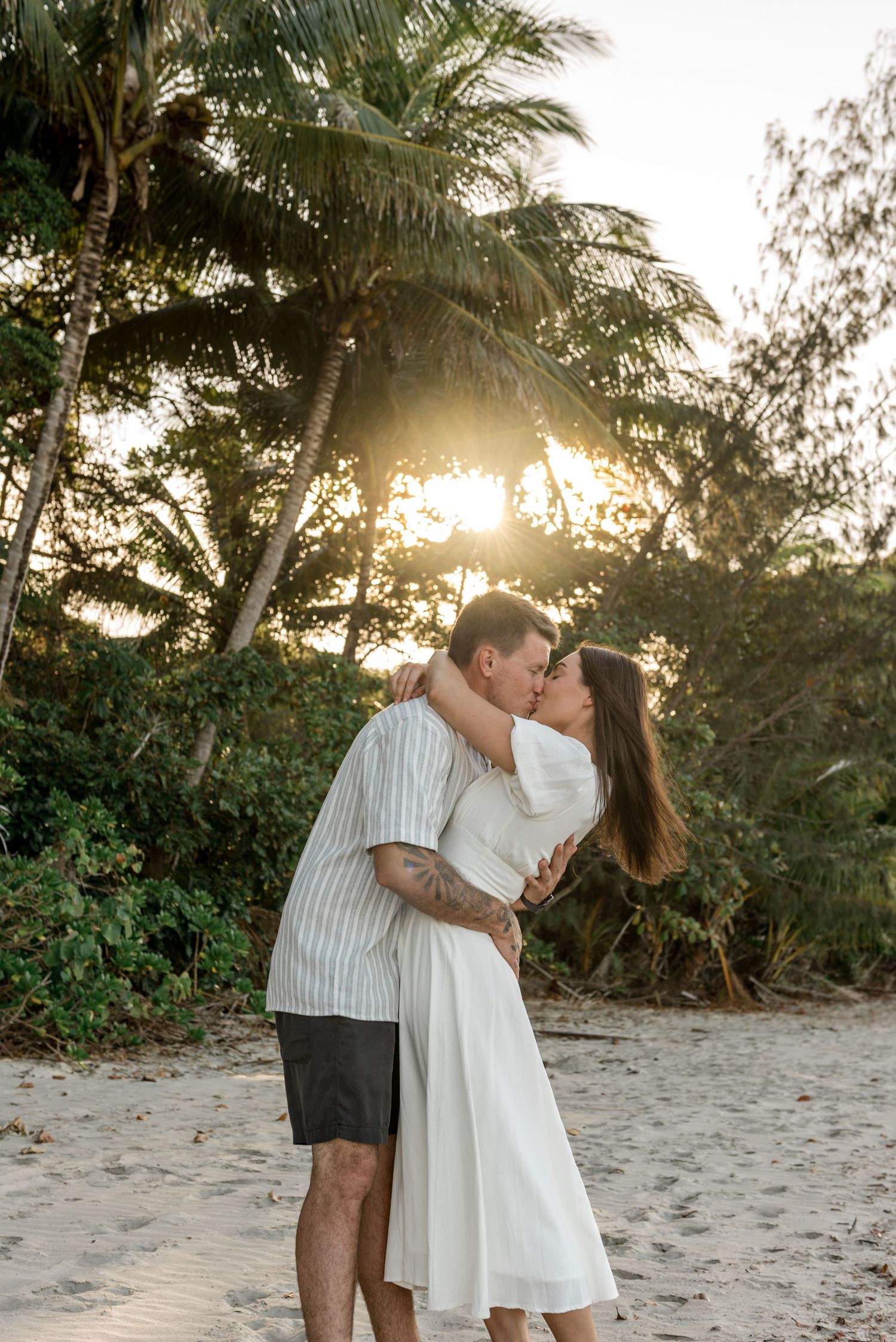 Couple sharing a romantic kiss on a sandy beach with palm trees and golden sunset light behind them.