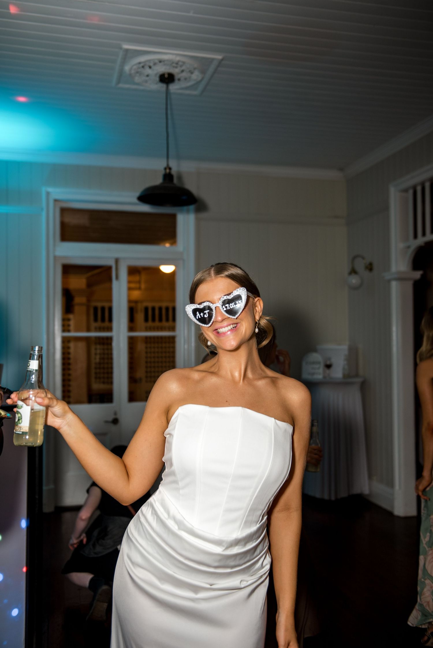 Bride in strapless white gown wearing heart-shaped novelty sunglasses holding a drink at her wedding reception.