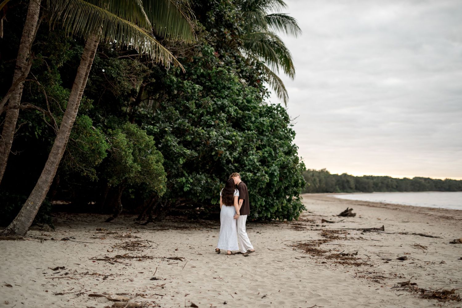 Couple embracing near lush tropical trees on a serene sandy beach under an overcast sky.