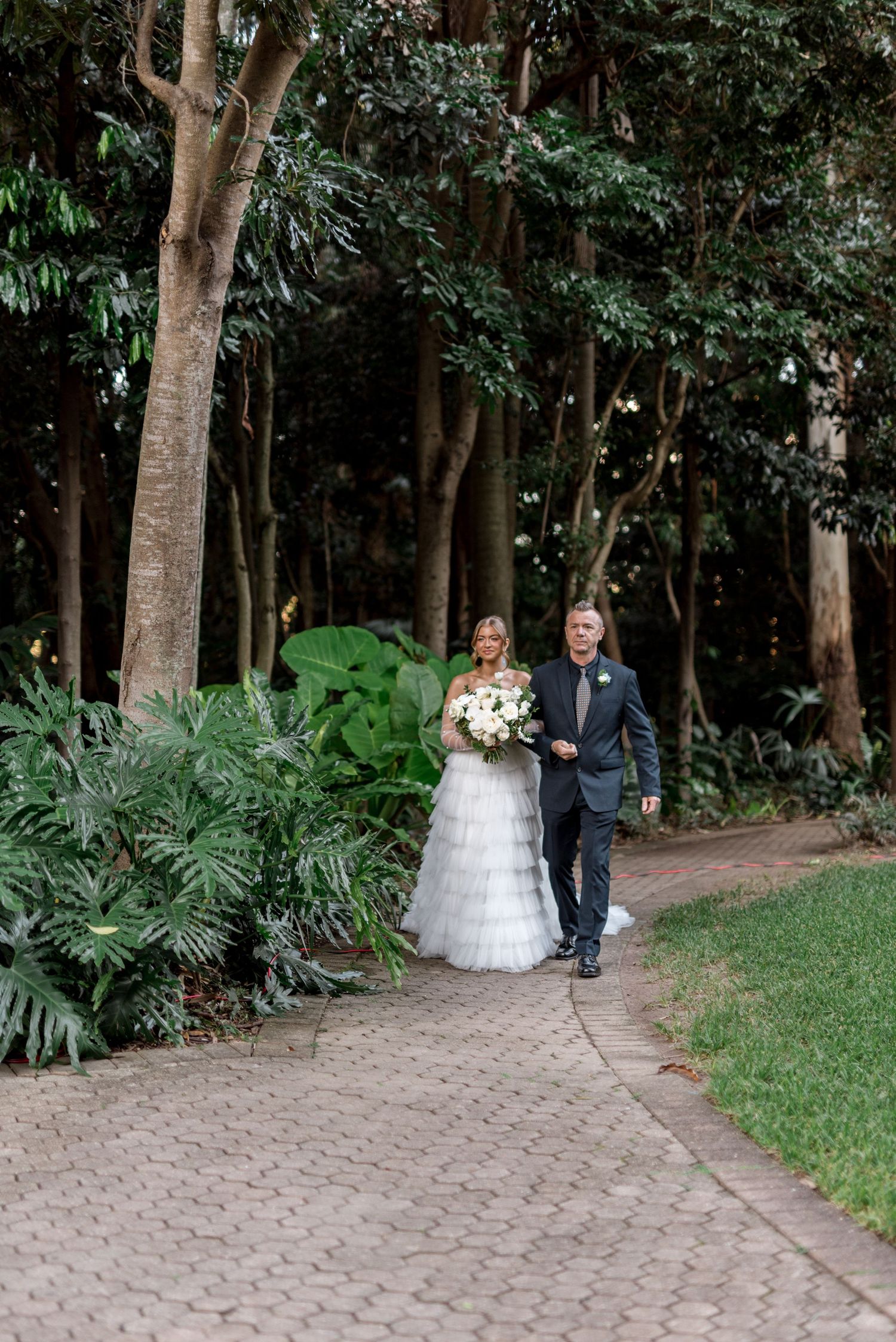 Bride in tiered tulle gown walks with escort along tree-lined cobblestone path to ceremony.
