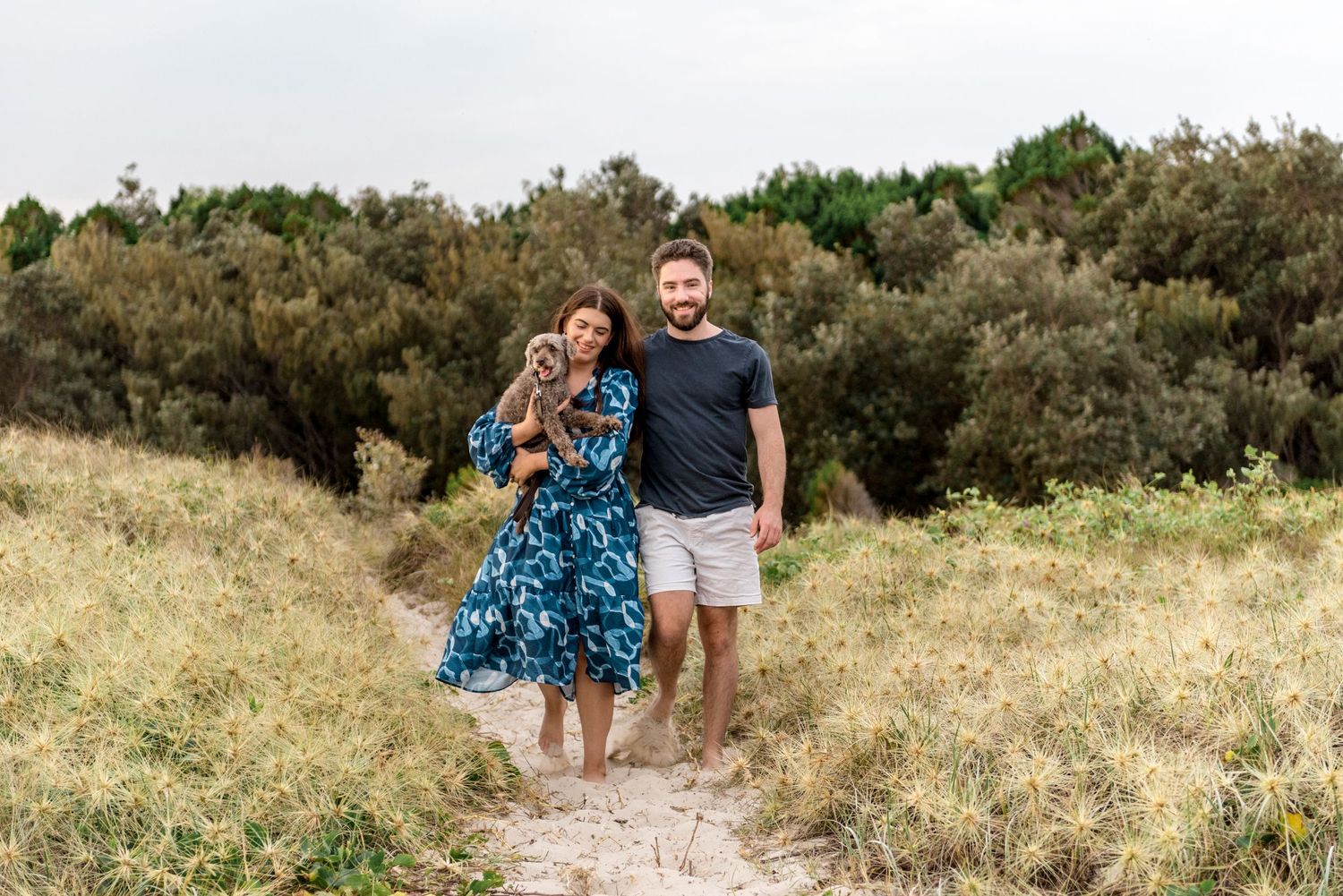Smiling couple standing on sandy path through beach dunes, woman holding gray dog in blue dress.