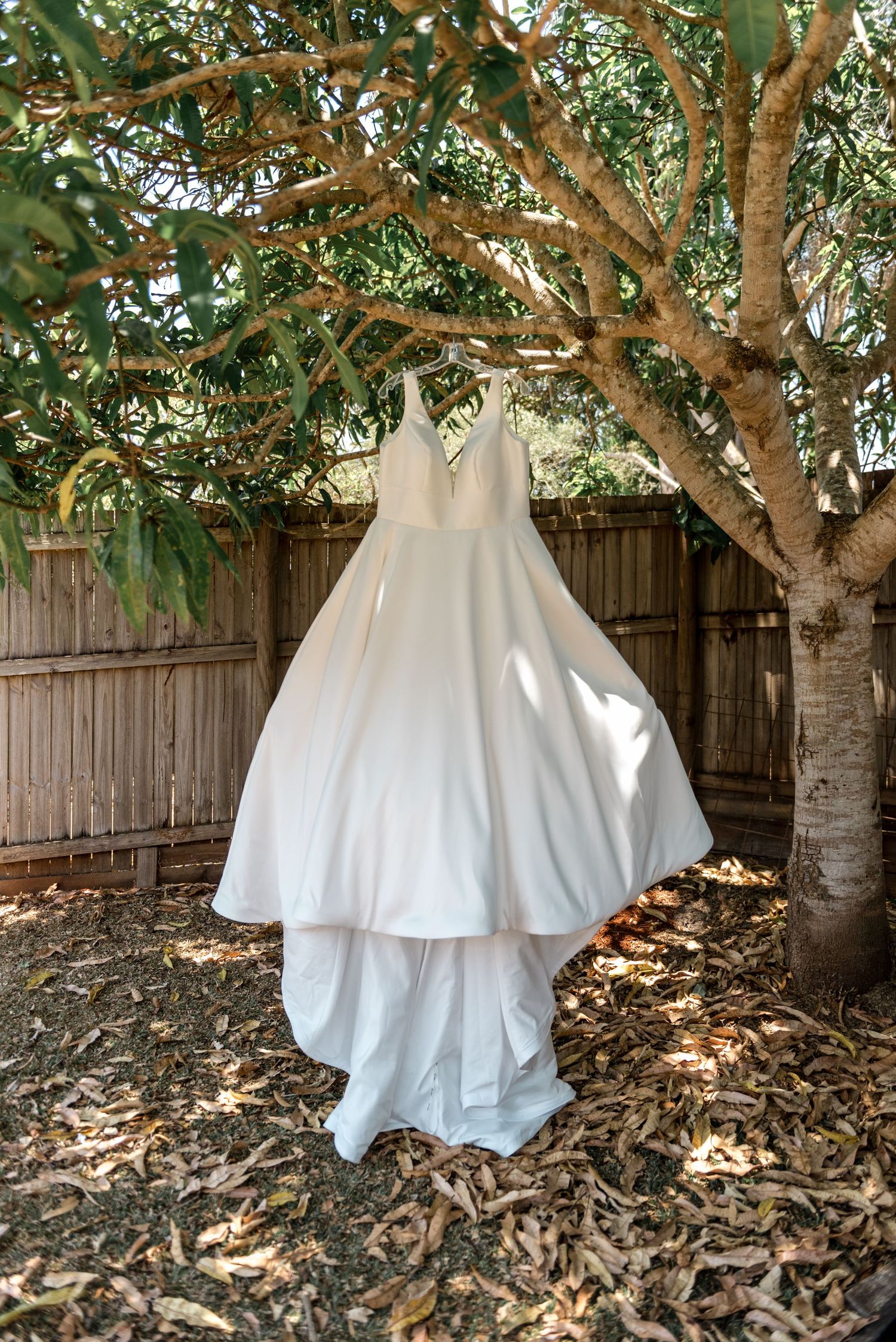 Beautiful white bridal gown with a full skirt hanging from a tree in a sunlit garden setting.