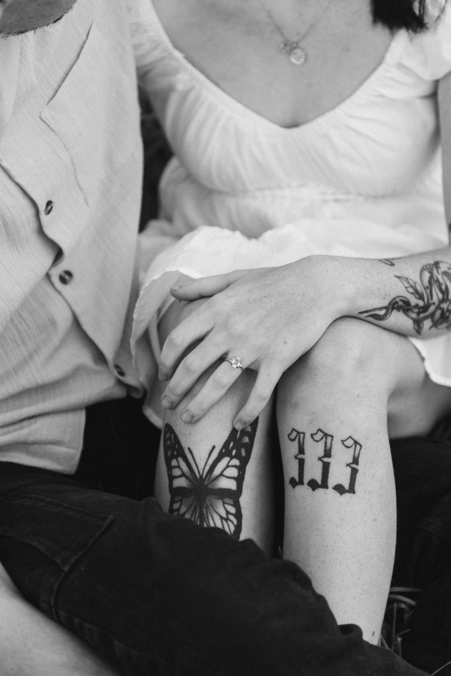 Black and white close-up of couple showing engagement ring beside butterfly and 333 tattoos.
