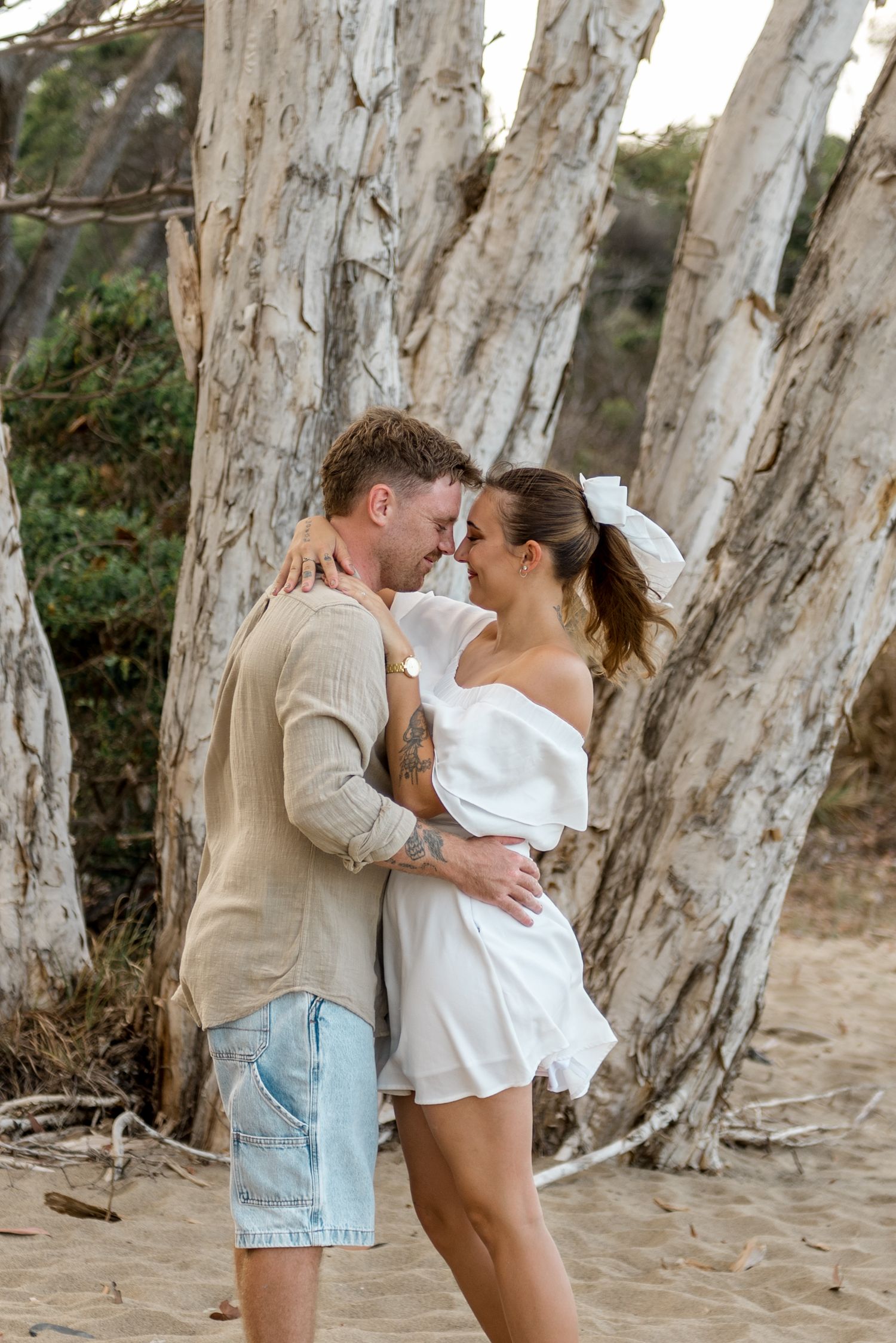 Couple embracing and almost kissing among white eucalyptus trees on a sandy beach.