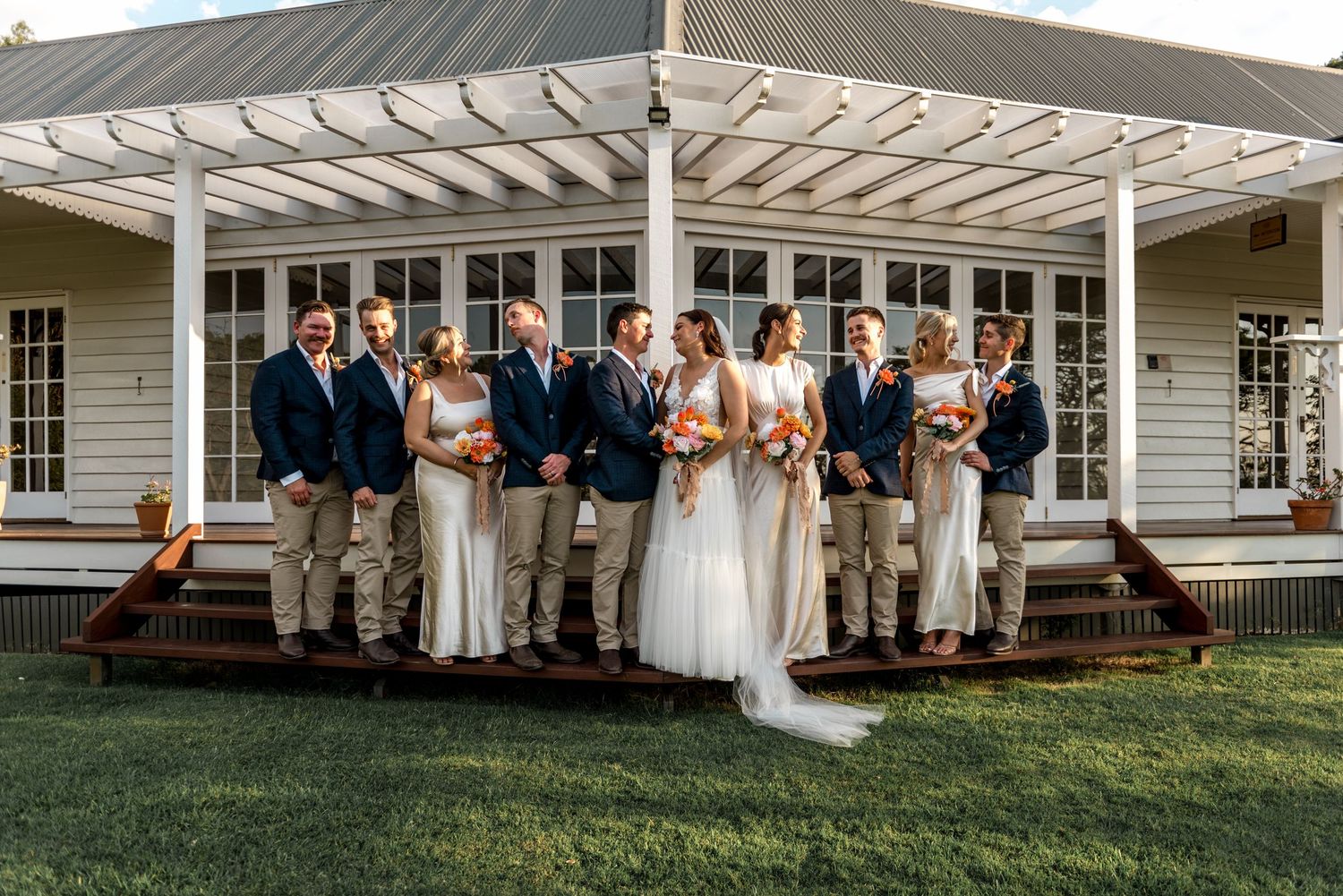 Wedding party posing on a white farmhouse porch with groomsmen in navy and bridesmaids in white.