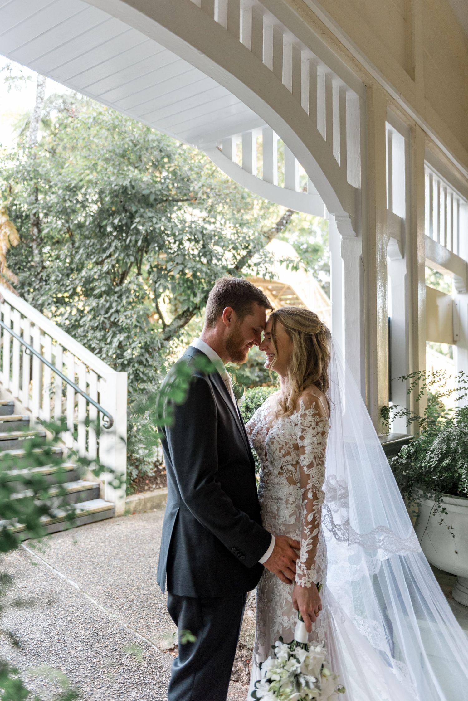 Bride and groom share an intimate forehead touch under a white arched veranda surrounded by greenery.