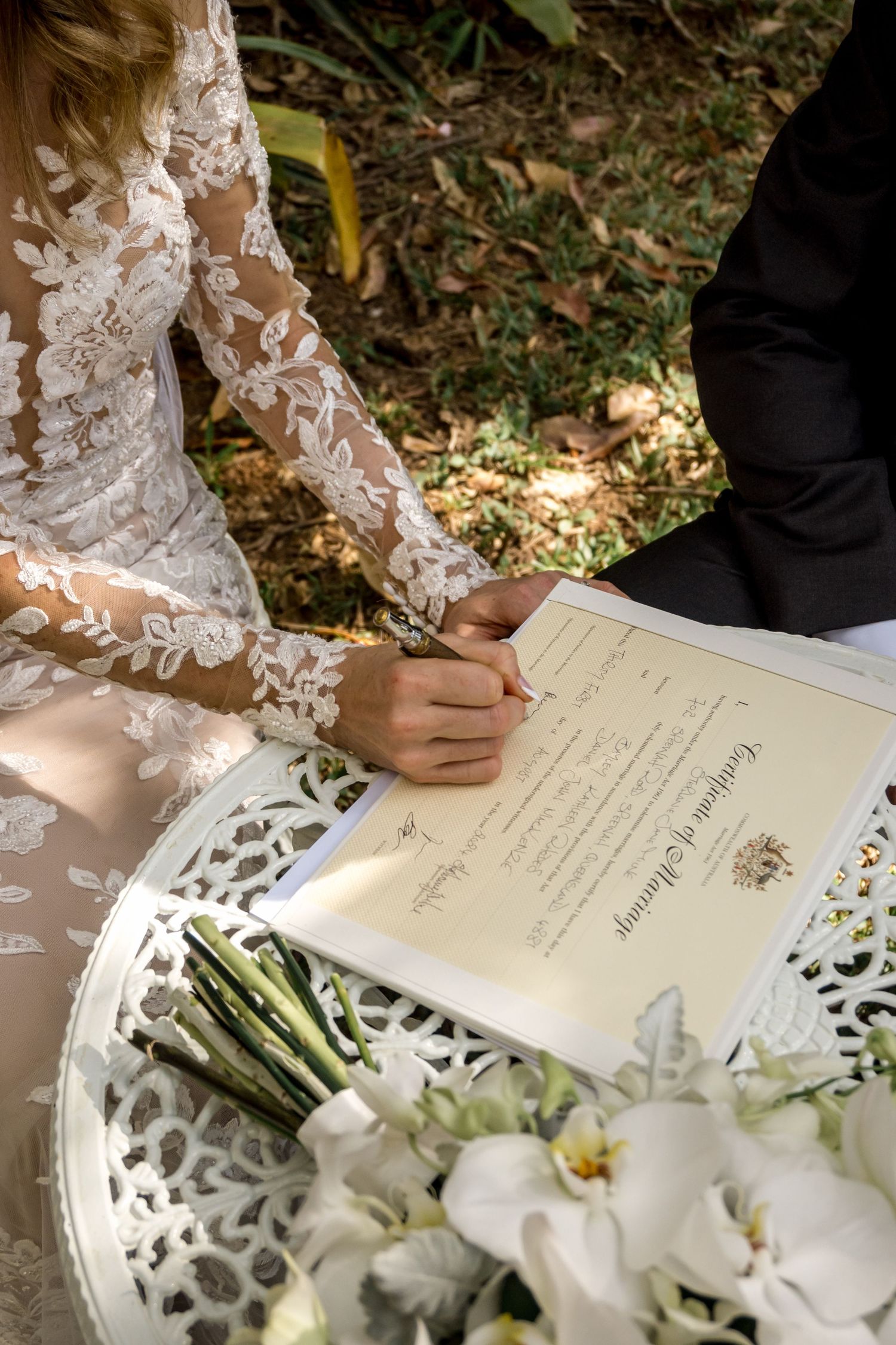 Bride signs marriage certificate on white table with white orchid bouquet nearby.