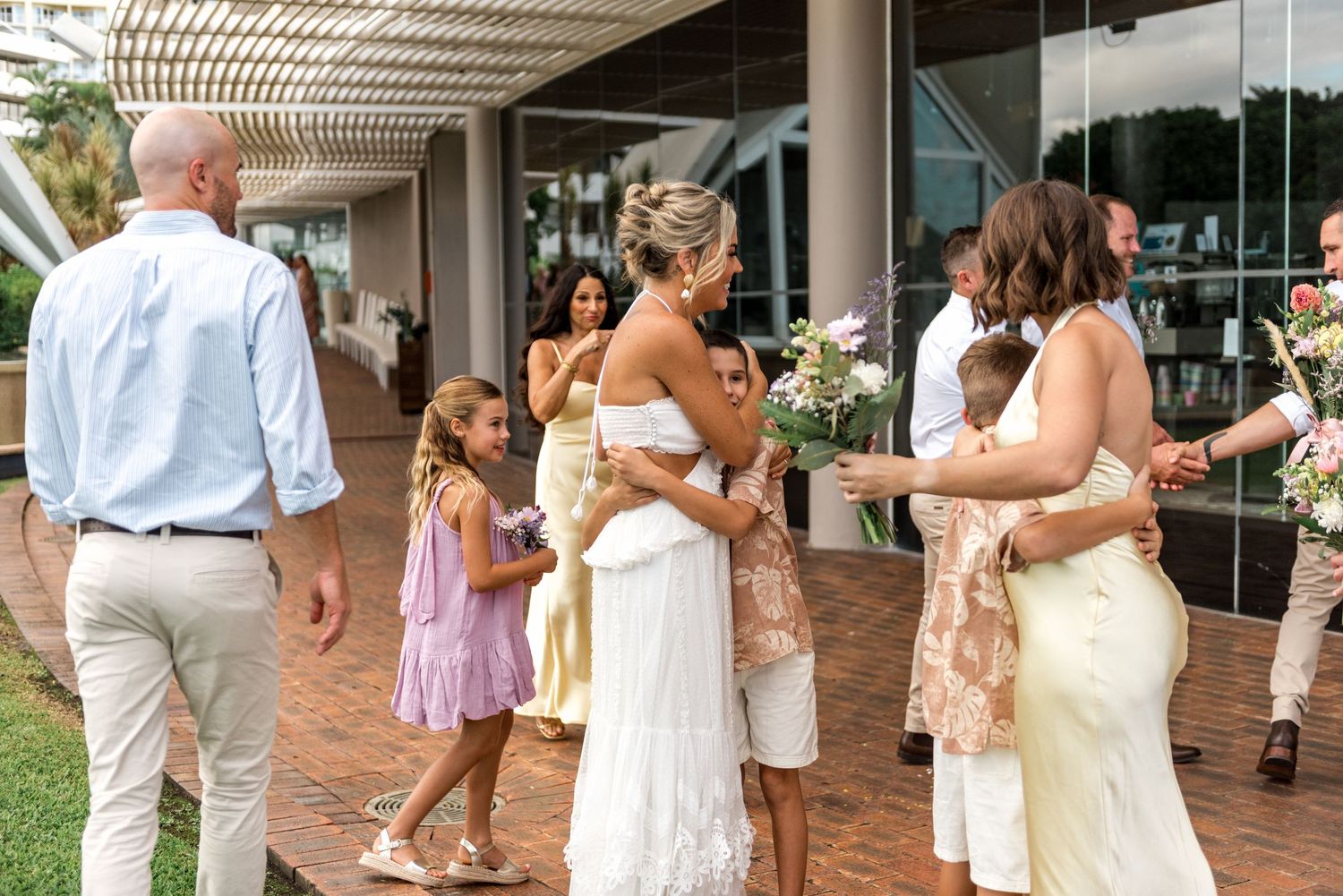 Wedding guests congratulate the bride in a white fringe dress outside a modern venue.