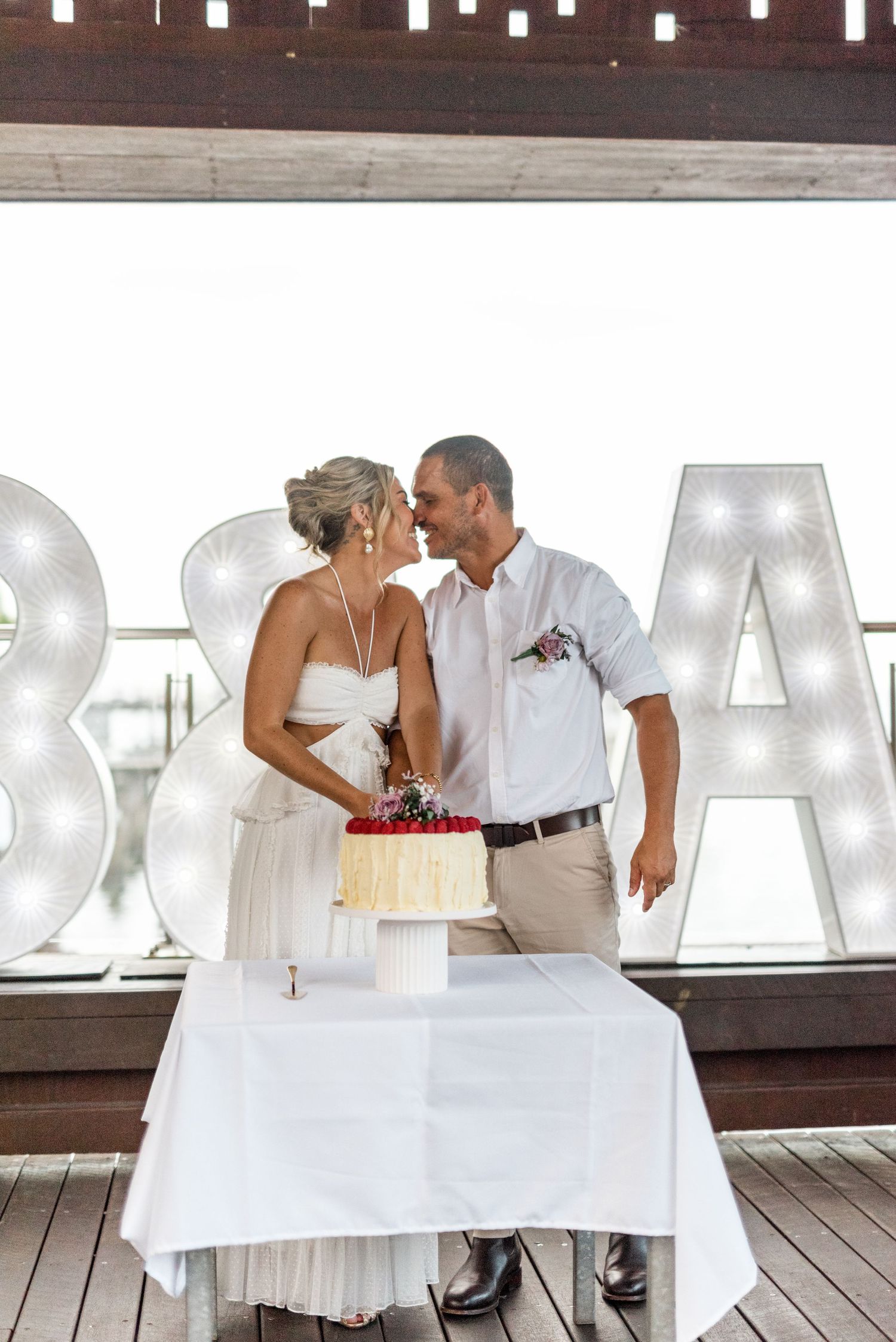 Bride and groom sharing romantic moment behind wedding cake with illuminated letter signs backdrop.