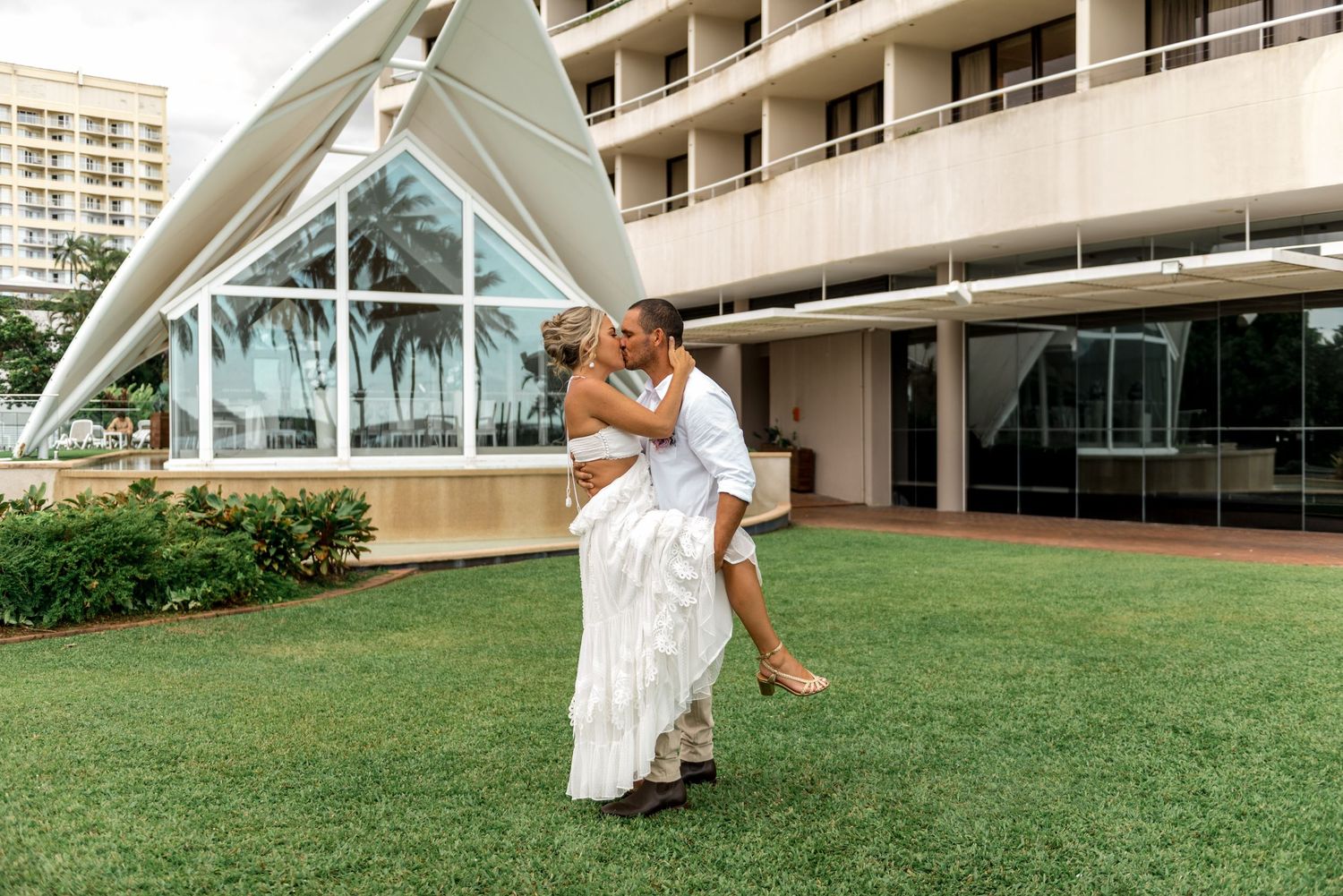 Groom lifts bride in passionate kiss on green lawn outside elegant hotel with glass architecture.