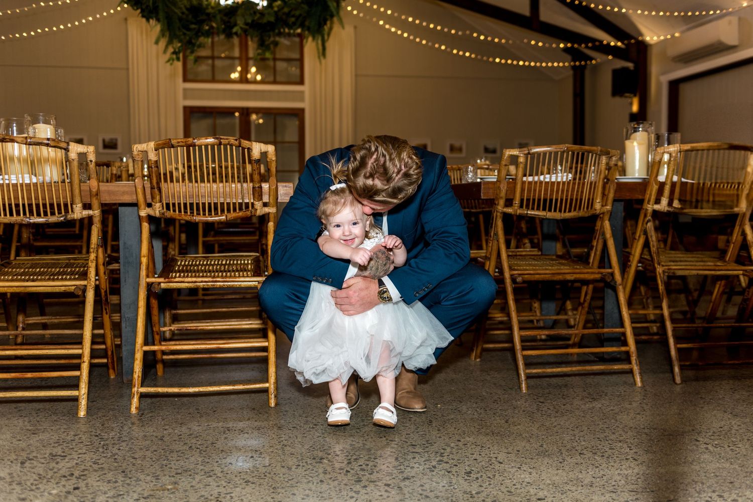 Groom in navy suit tenderly embracing a small flower girl in a white tutu dress at a wedding reception.