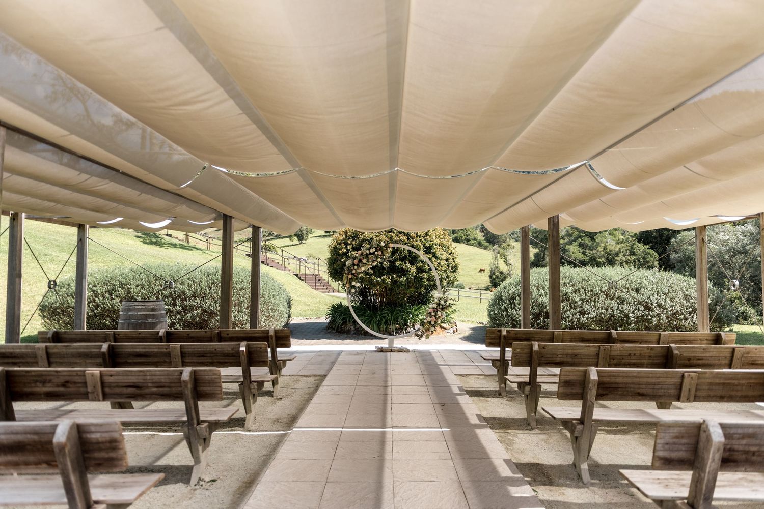 Outdoor wedding ceremony venue with wooden pews under shade sails and garden backdrop.